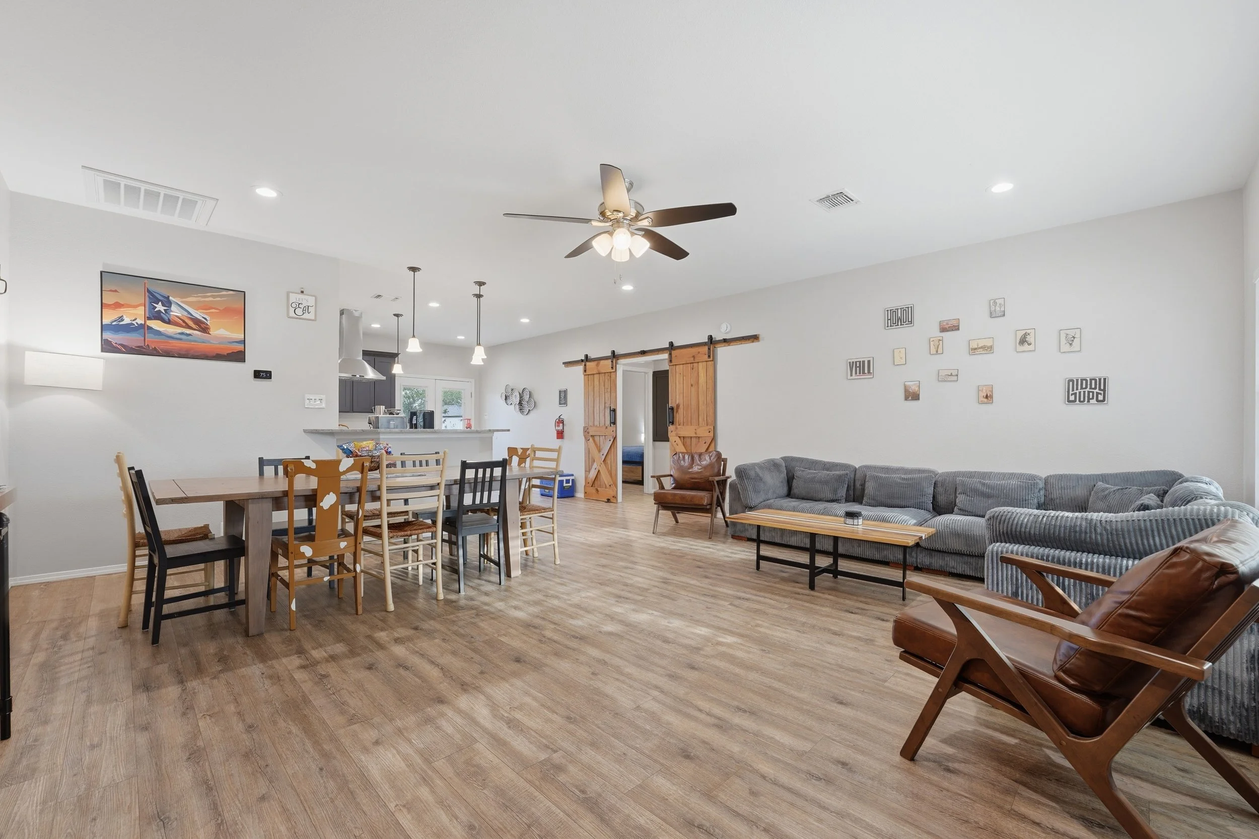 Open-concept living room and kitchen with white walls, wooden flooring, ceiling fan, and various seating options.