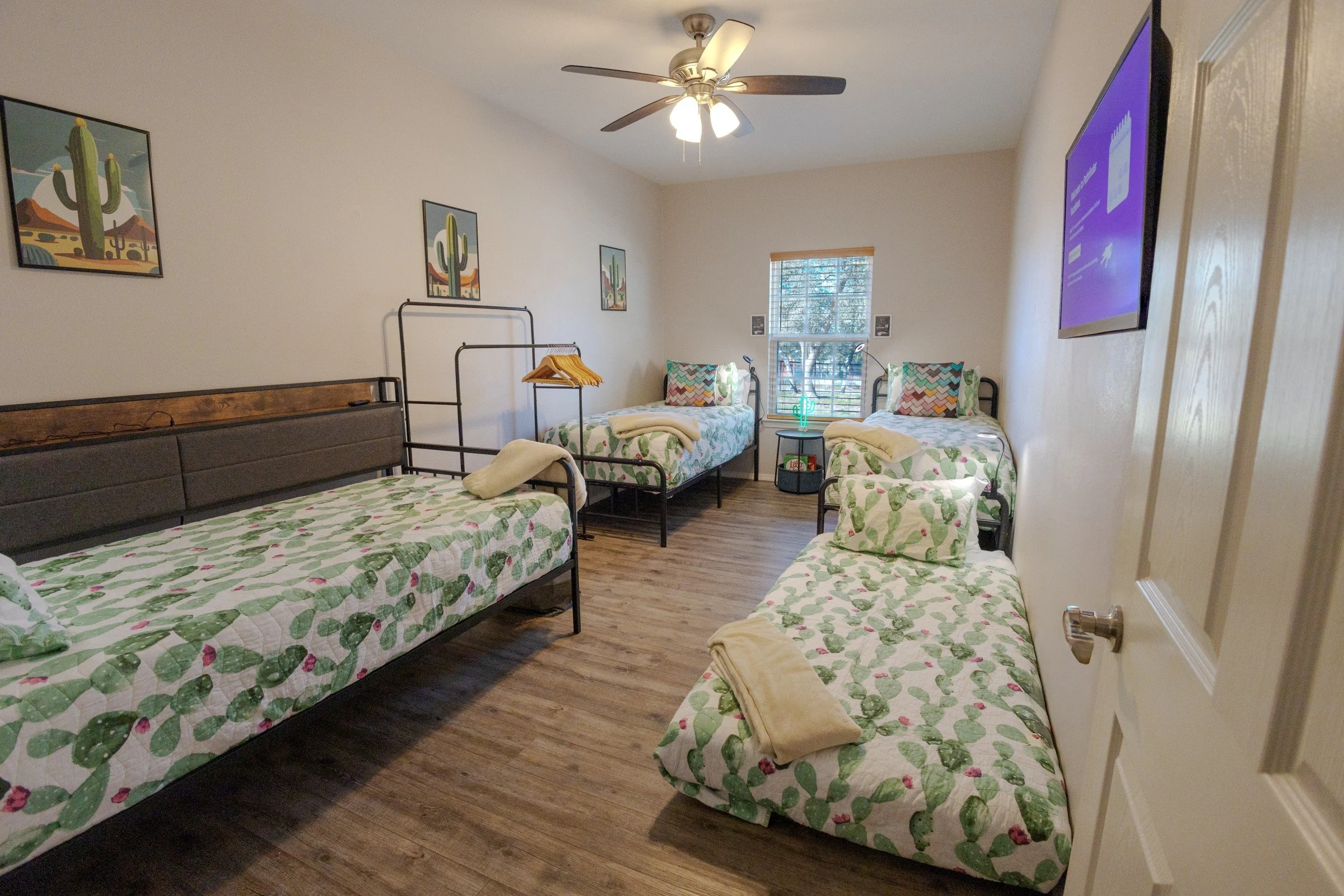 A bedroom with five beds covered in cactus-themed bedspreads, each with a towel placed on top. There are three framed cactus art prints on the wall, a ceiling fan, a window with blinds, and a small black side table with a neon cactus light and books.