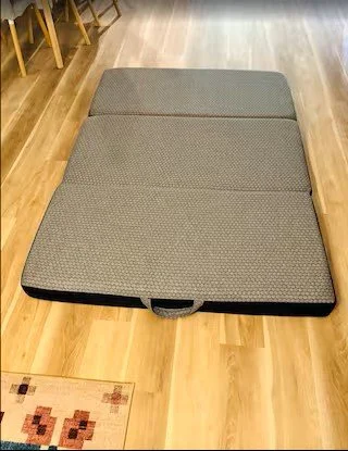 Foldable foam mattress on a wooden floor under a dining table.