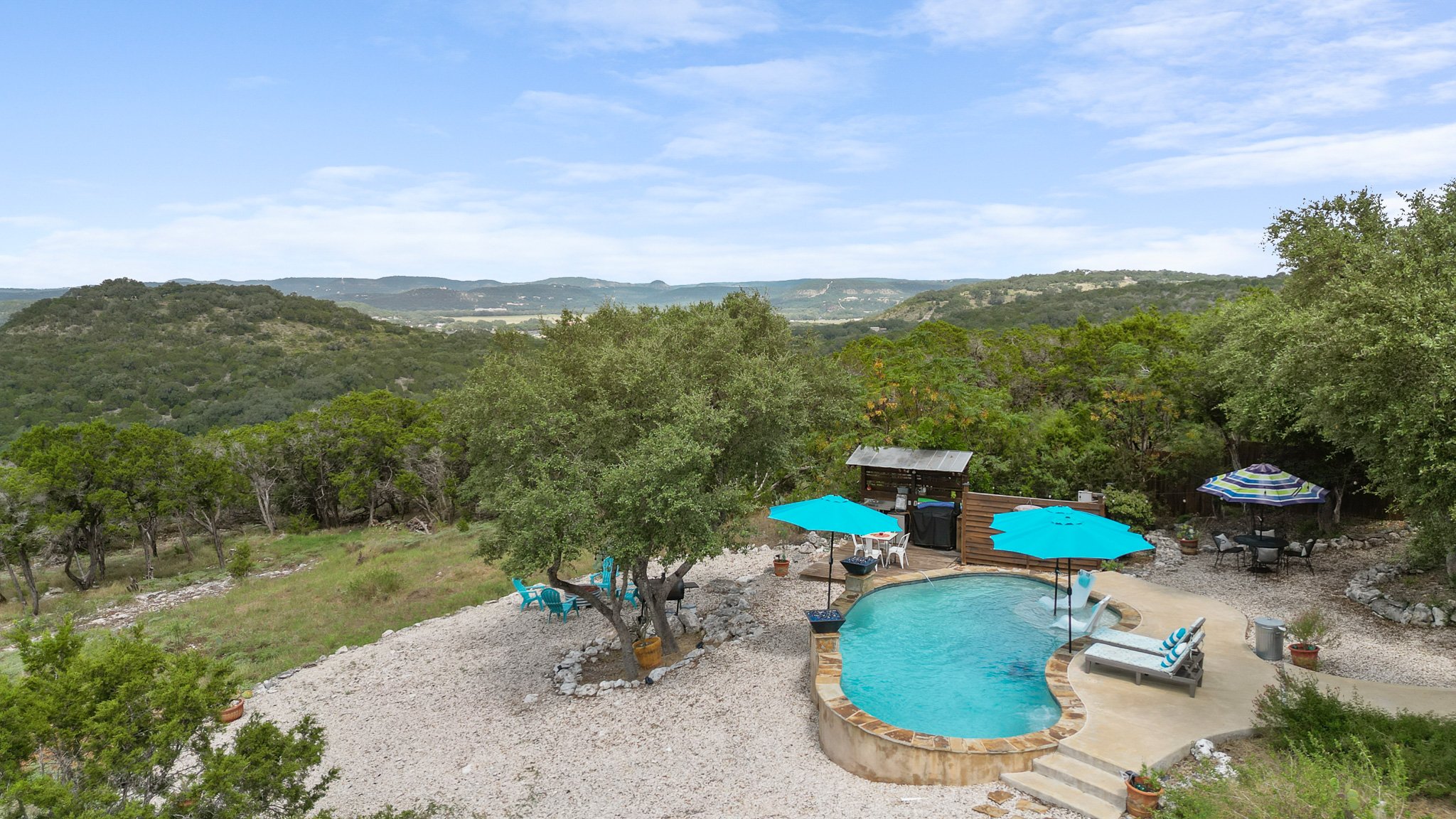 Backyard with a swimming pool, outdoor seating, umbrellas, and lush green hills in the background.