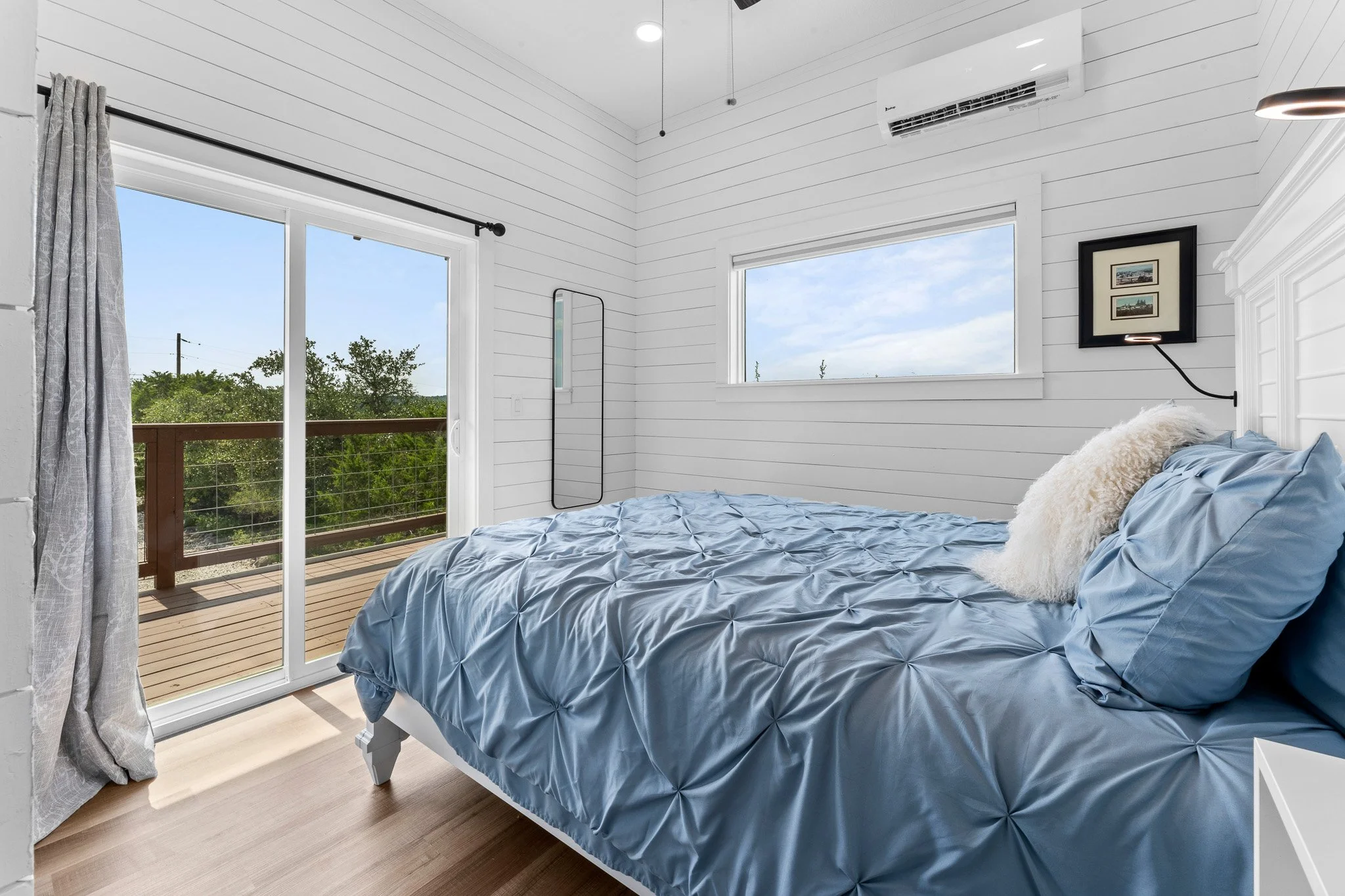 A bedroom with white shiplap walls, a bed with blue ruched bedding and fluffy pillow, large window and sliding glass door leading to balcony, tree view outside, framed pictures on wall, air conditioning unit, and wooden flooring.