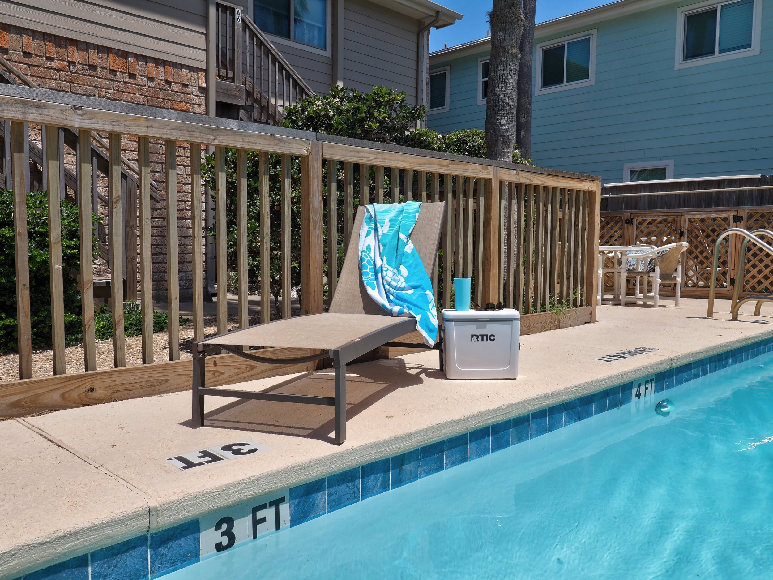 An empty backyard swimming pool with a light beige concrete deck. On the edge of the pool is a lounge chair with a turquoise towel draped over it. Next to the lounge chair is a small cooler and a blue cup. The pool's depth markings indicate 3 and 4 f