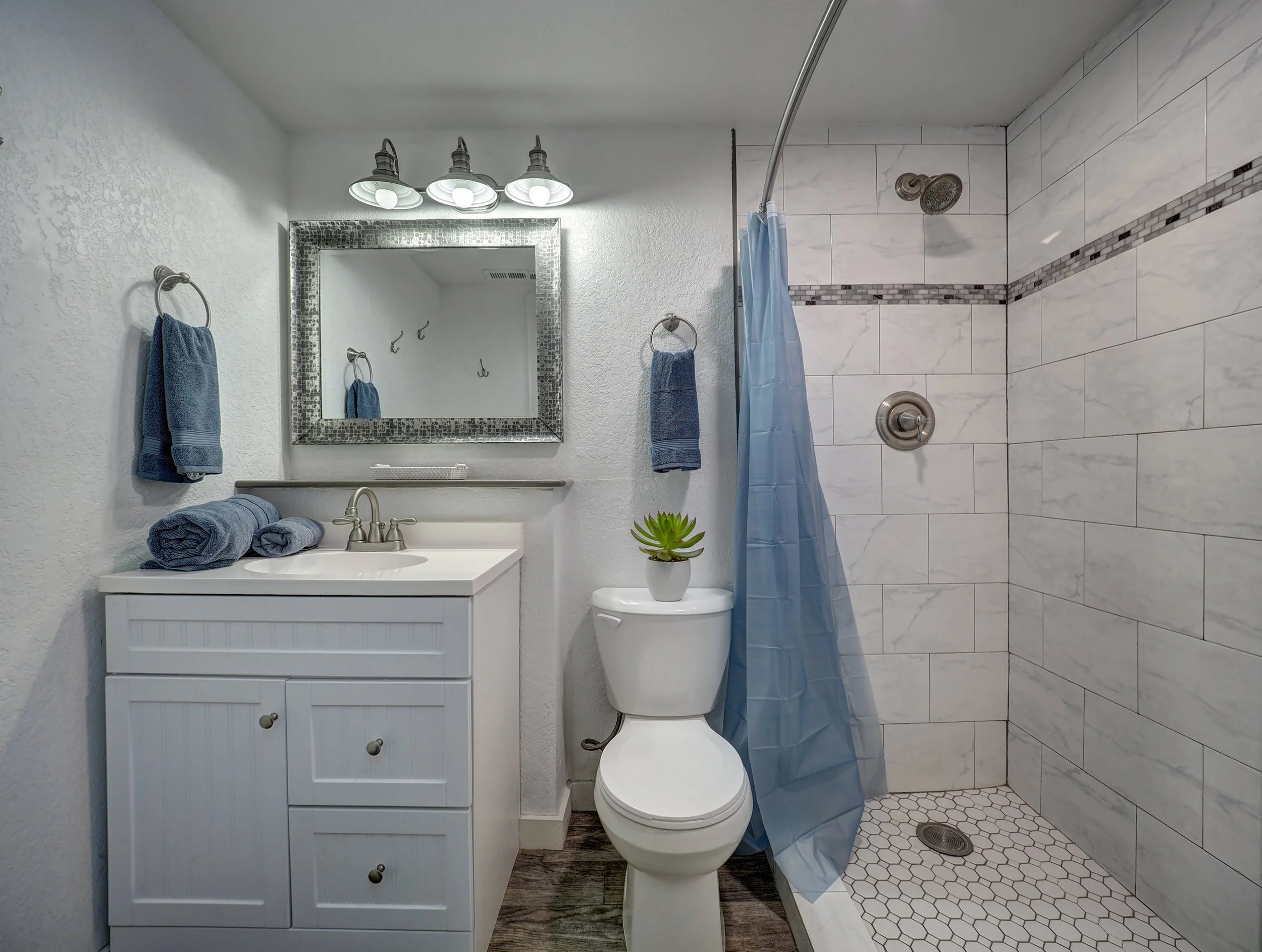 A small bathroom with a white vanity, a mirror above it, and a toilet. Towels hang on the wall, and there is a plant on the toilet tank. The shower area has white marble tiles with a decorative strip and a blue shower curtain. The floor has hexagonal