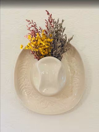 A white vase with dried flowers, including yellow and purple sprigs, placed on a decorative white plate on a white surface.