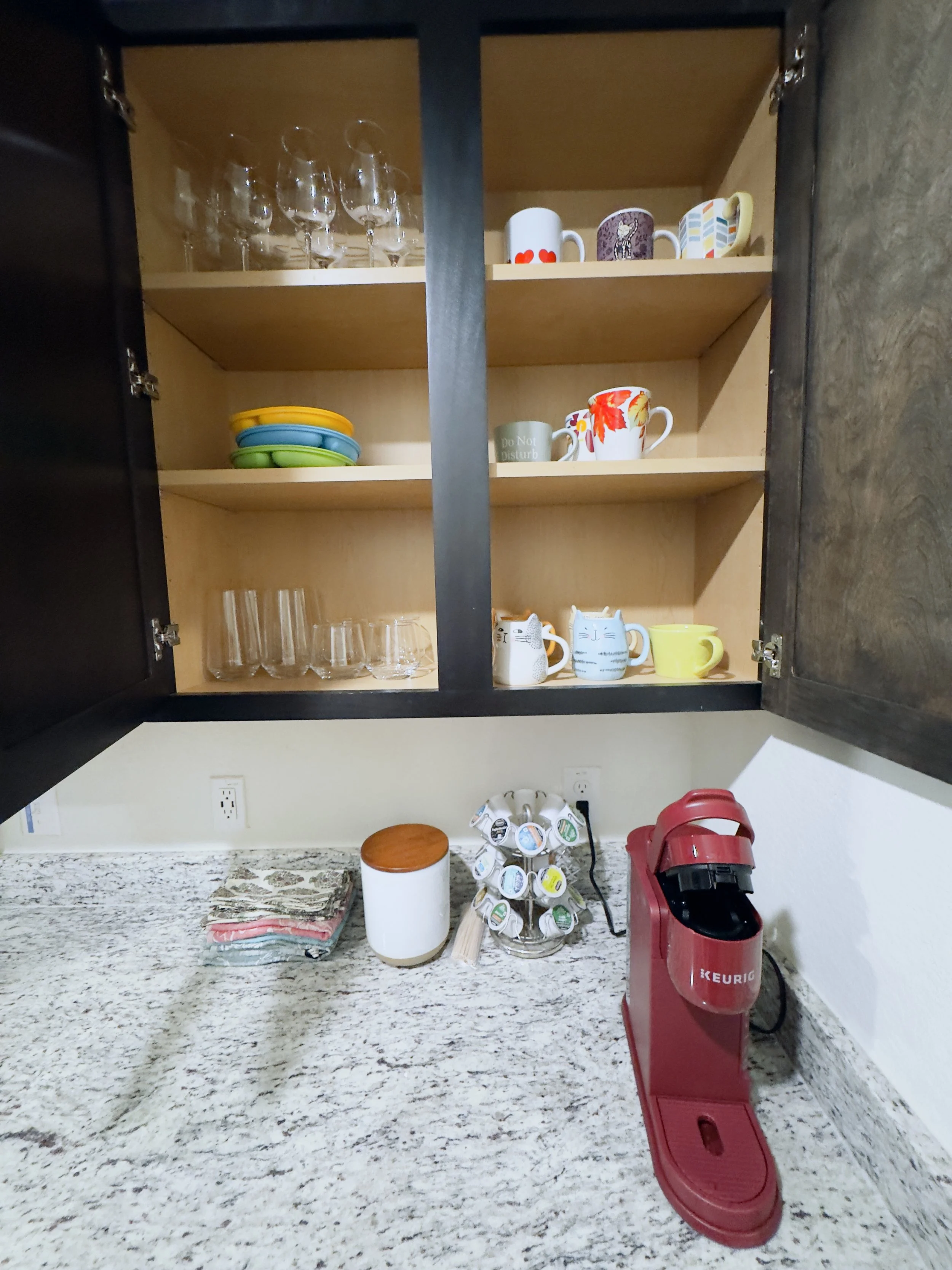 Open kitchen cabinet with glasses on top shelf, coffee mugs and bowls on middle shelf, counter with dish towel, coffee pod holder, red Keurig coffee machine, and a container on the countertop.
