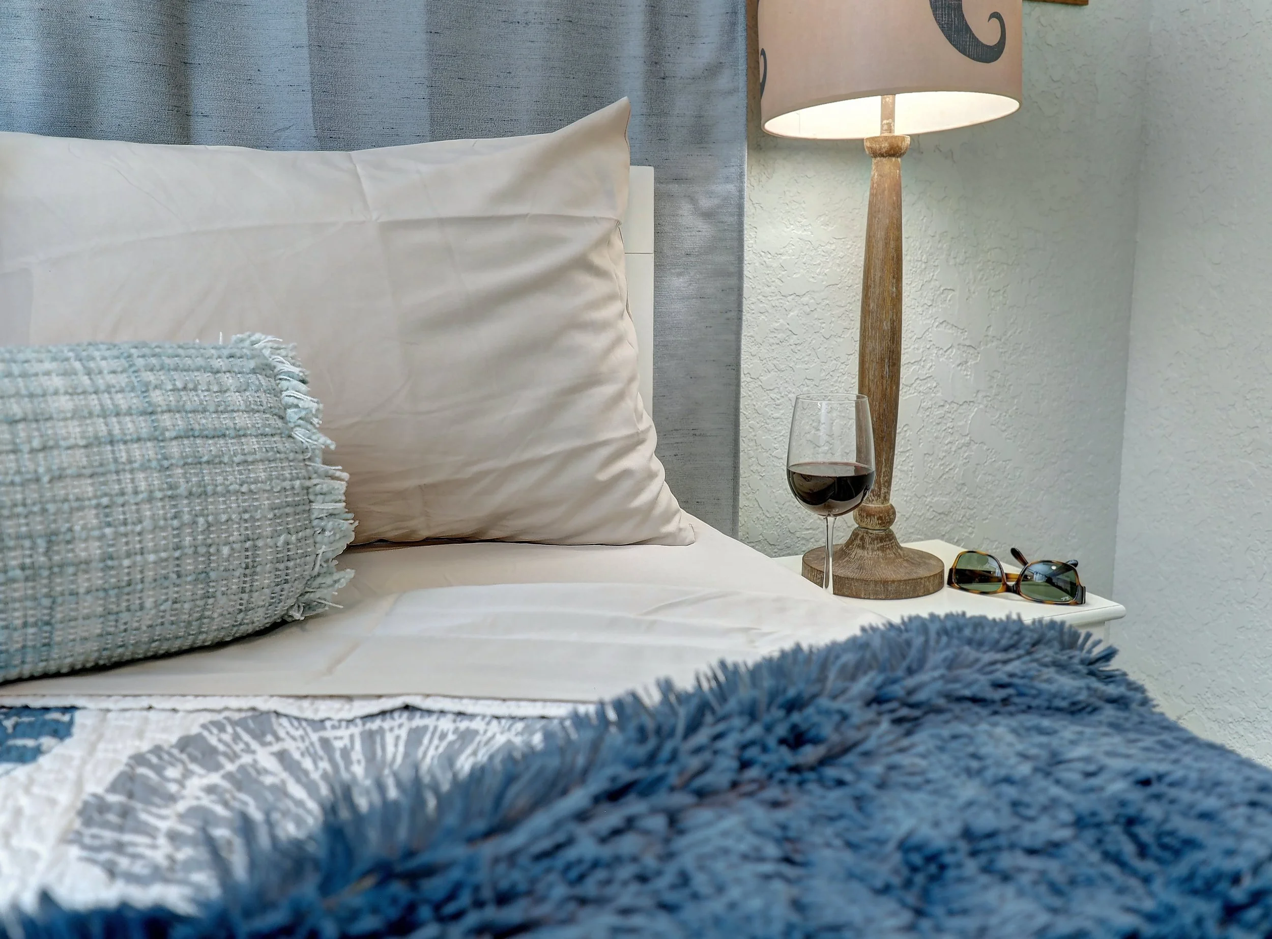 A close-up of a bed with white pillows, a gray woven blanket, and a blue fringed throw; a window with a textured curtain, a wooden bedside lamp, a glass of red wine, sunglasses, and part of a blue and white patterned bedspread.