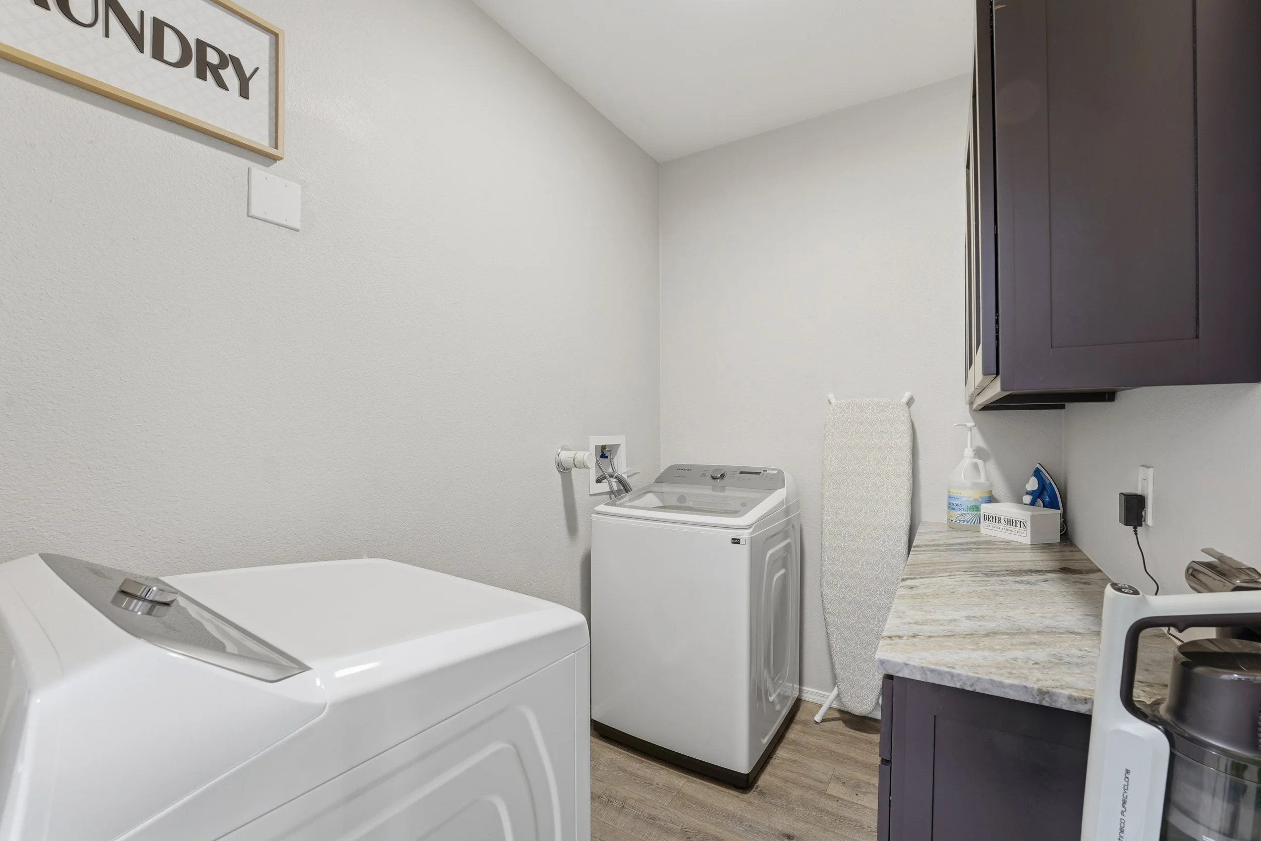 A laundry room with a white top-loading washing machine, a white top-loading dryer, a dark purple cabinet, an ironing board, and laundry supplies on a countertop.