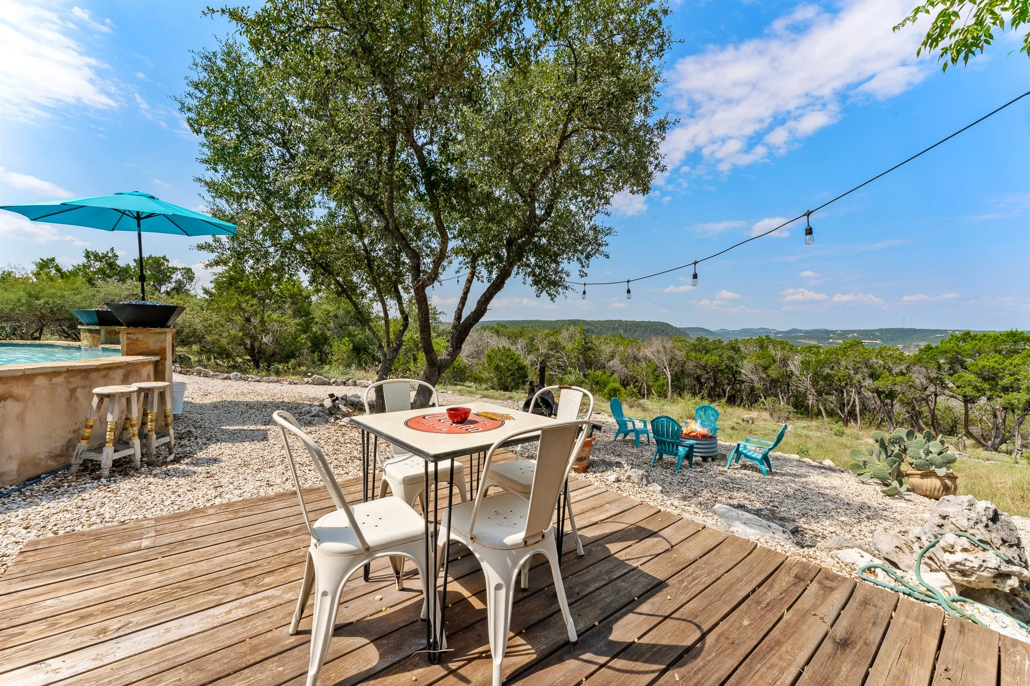 Outdoor patio with white plastic chairs around a table with a red bowl, on a wooden deck, with a tree and string lights in a rural landscape under a partly cloudy sky.