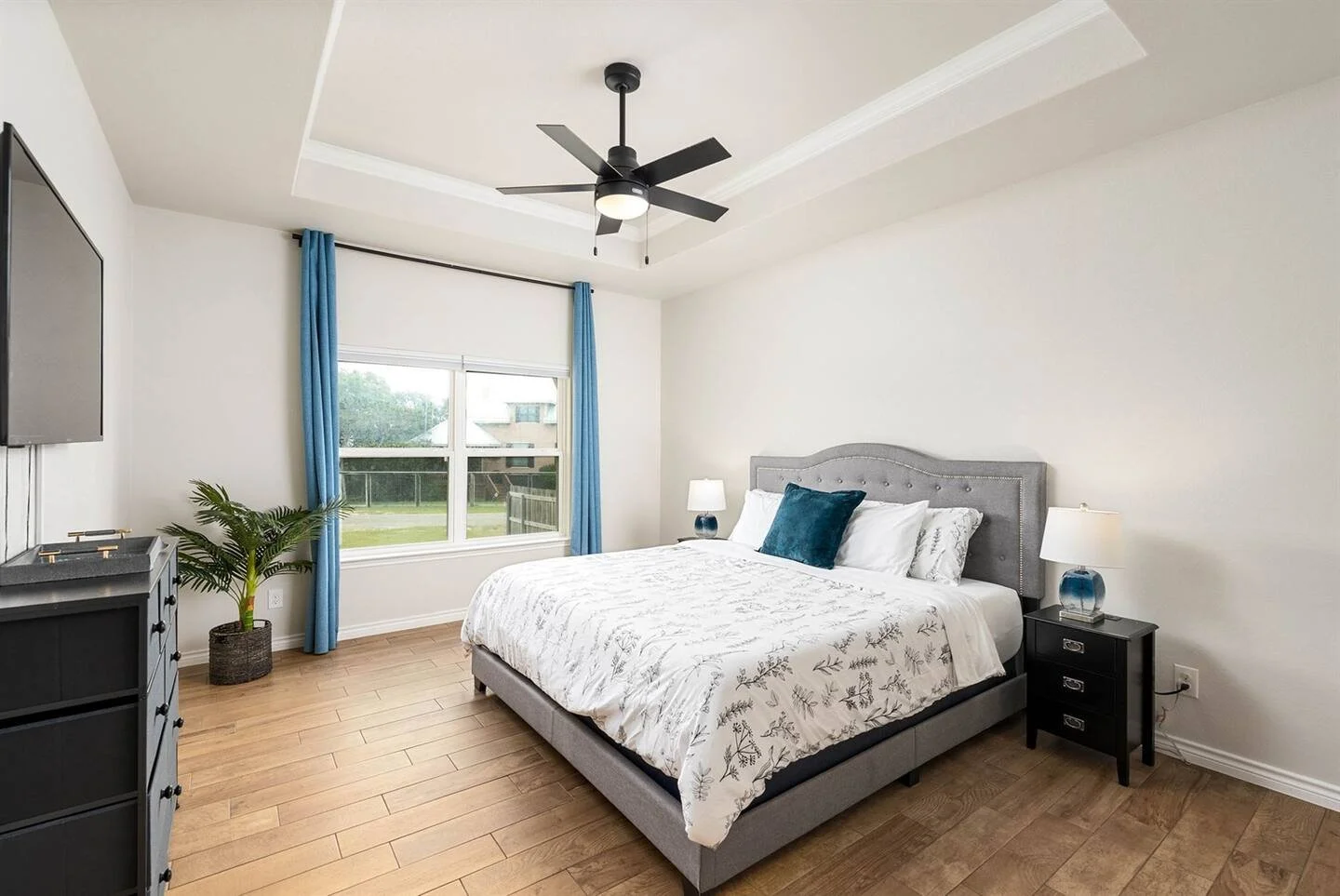 Modern bedroom with a queen bed, gray upholstered headboard, white bedding with leaf patterns, blue pillows, black nightstand with a lamp, a dresser with a TV, a ceiling fan, blue curtains, window showing a view of a grassy yard.