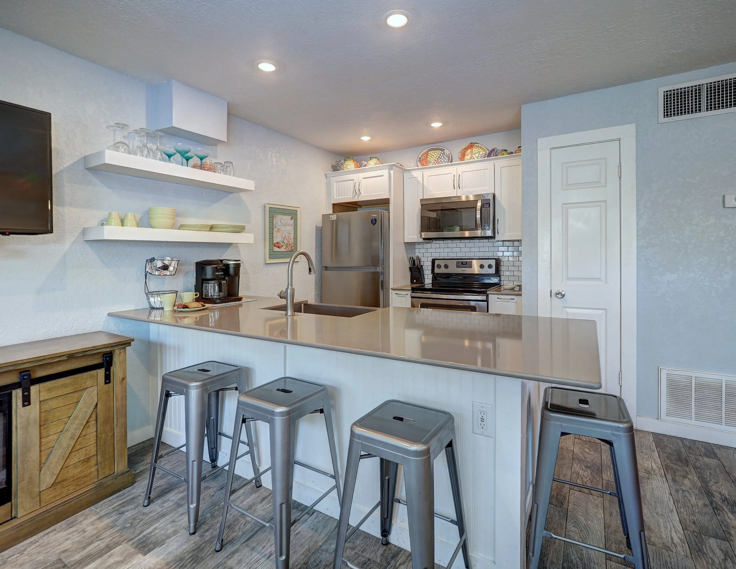 Kitchen with white cabinets, stainless steel appliances, and open shelves with dishes, glasses, and decorative items. Four metal stools are positioned around a kitchen island with a gray countertop, and a wooden cabinet and floor are also visible.