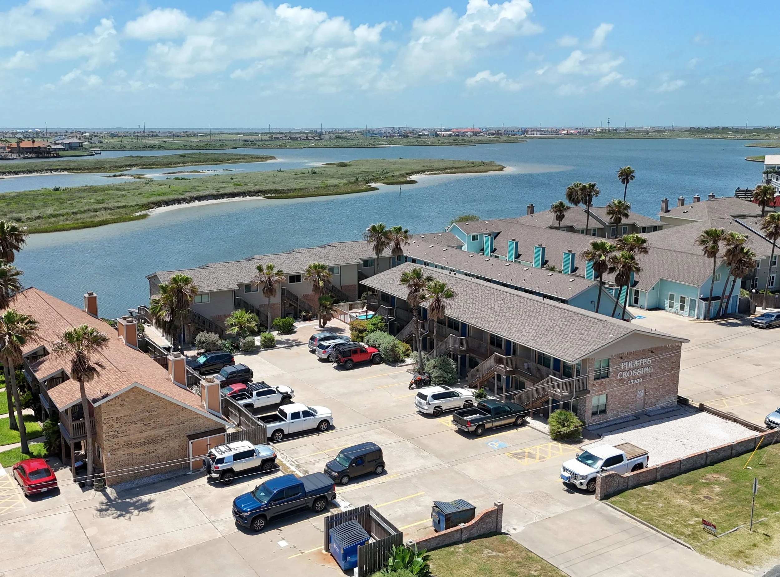 A multi-story apartment complex next to a waterway with beachfront houses in the background, parked cars in the foreground, and a partly cloudy sky.