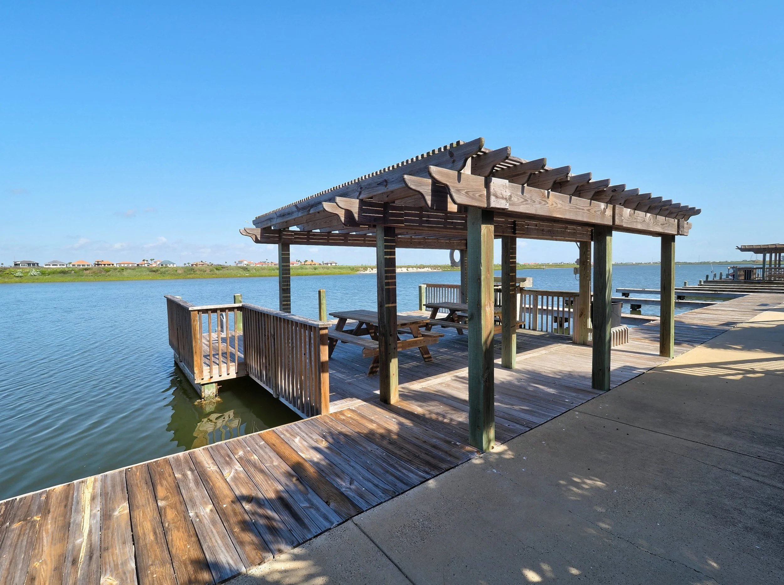 Wooden deck with a covered seating area by a body of water under a clear blue sky, with houses visible in the distance.