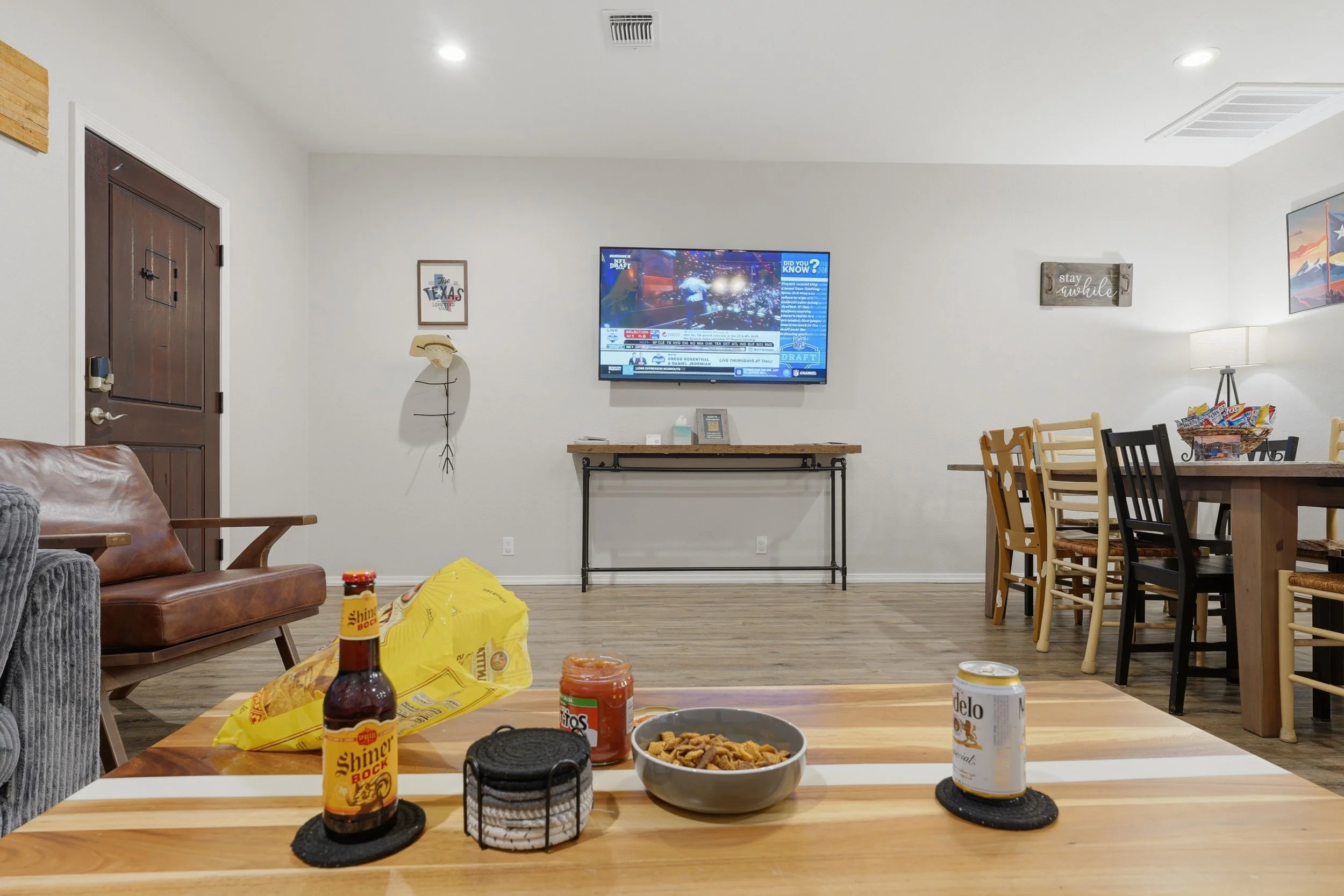 Living room with a wall-mounted TV, a wooden dining table with snacks and beverages, and a brown leather couch with a blanket, decorated with framed artwork and signs on the wall.