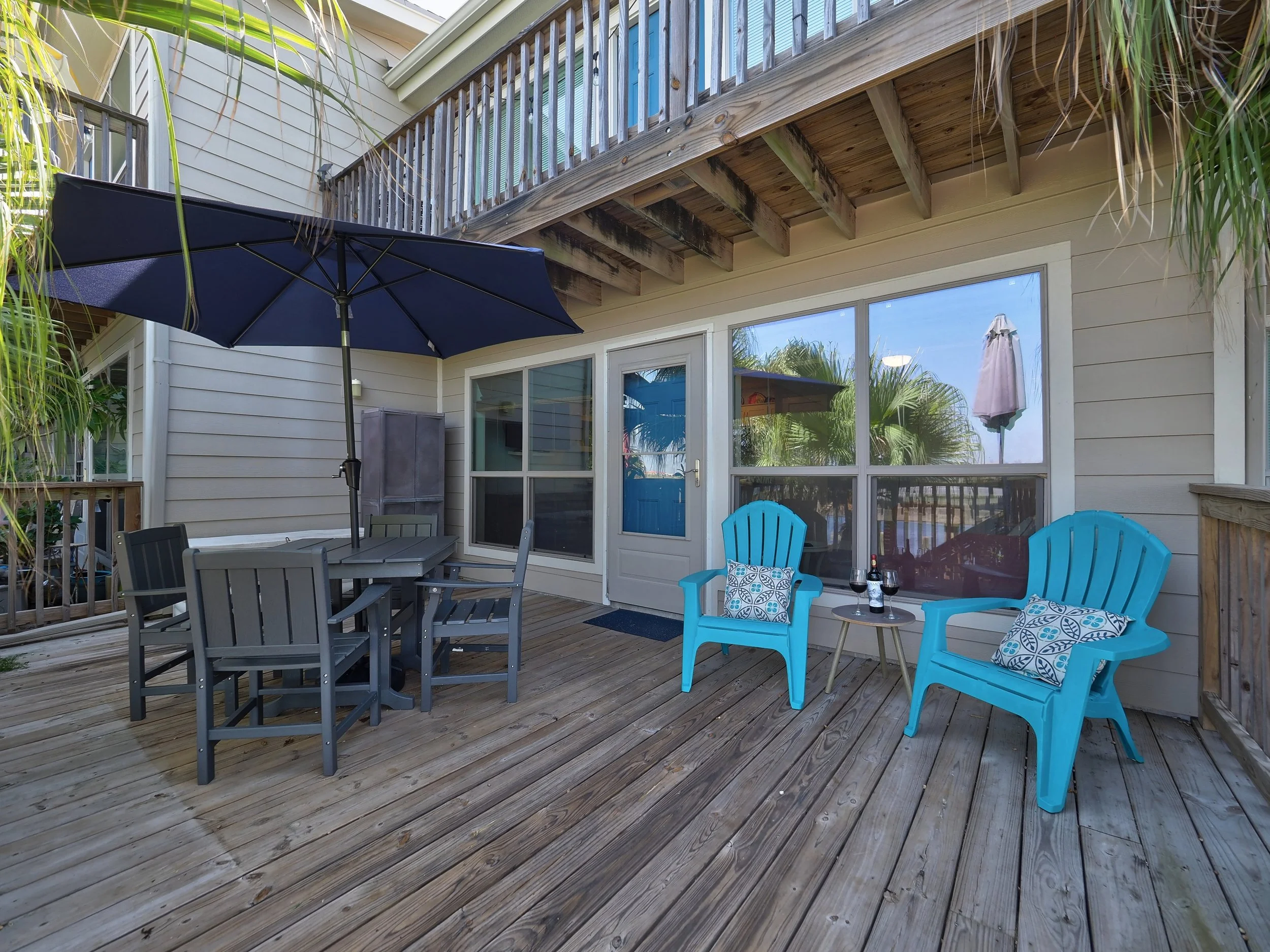 A backyard patio with wooden decking, outdoor furniture including a gray table with chairs, two bright blue plastic chairs, and a blue umbrella. There is a house with large windows and a door, and a balcony with wooden railing above.
