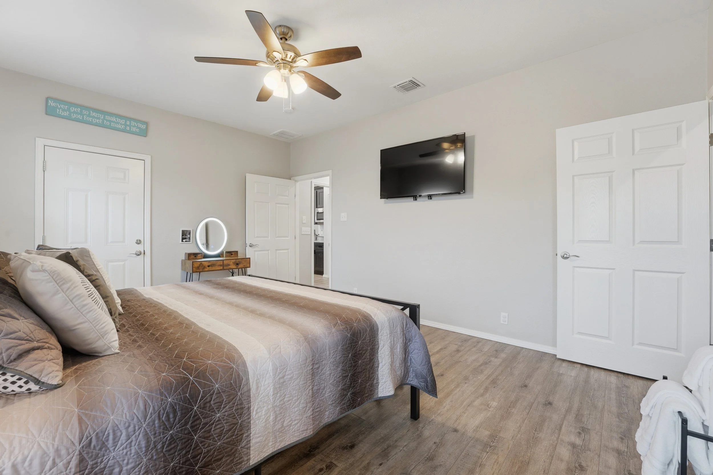 A bedroom with a large bed covered in a brown geometric patterned comforter and multiple pillows, a wooden dresser with a mirror, a mounted flat-screen TV, a ceiling fan with lights, and open white doors, with a sign on the wall that reads "Never get