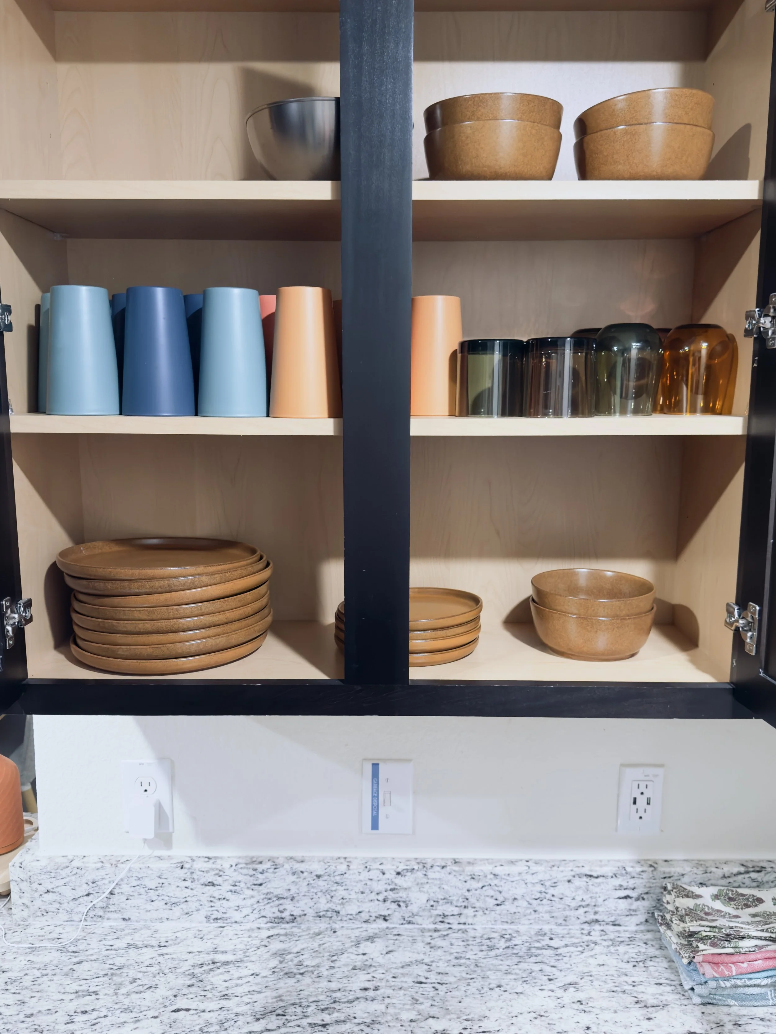 Open kitchen cabinet with two shelves displaying bowls, cups, and plates, above a granite countertop with folded napkins, and two electrical outlets on the wall.