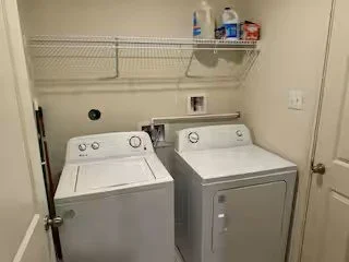 A laundry room with a white washing machine and a white dryer side by side, against a beige wall with a wire shelf above holding laundry supplies.