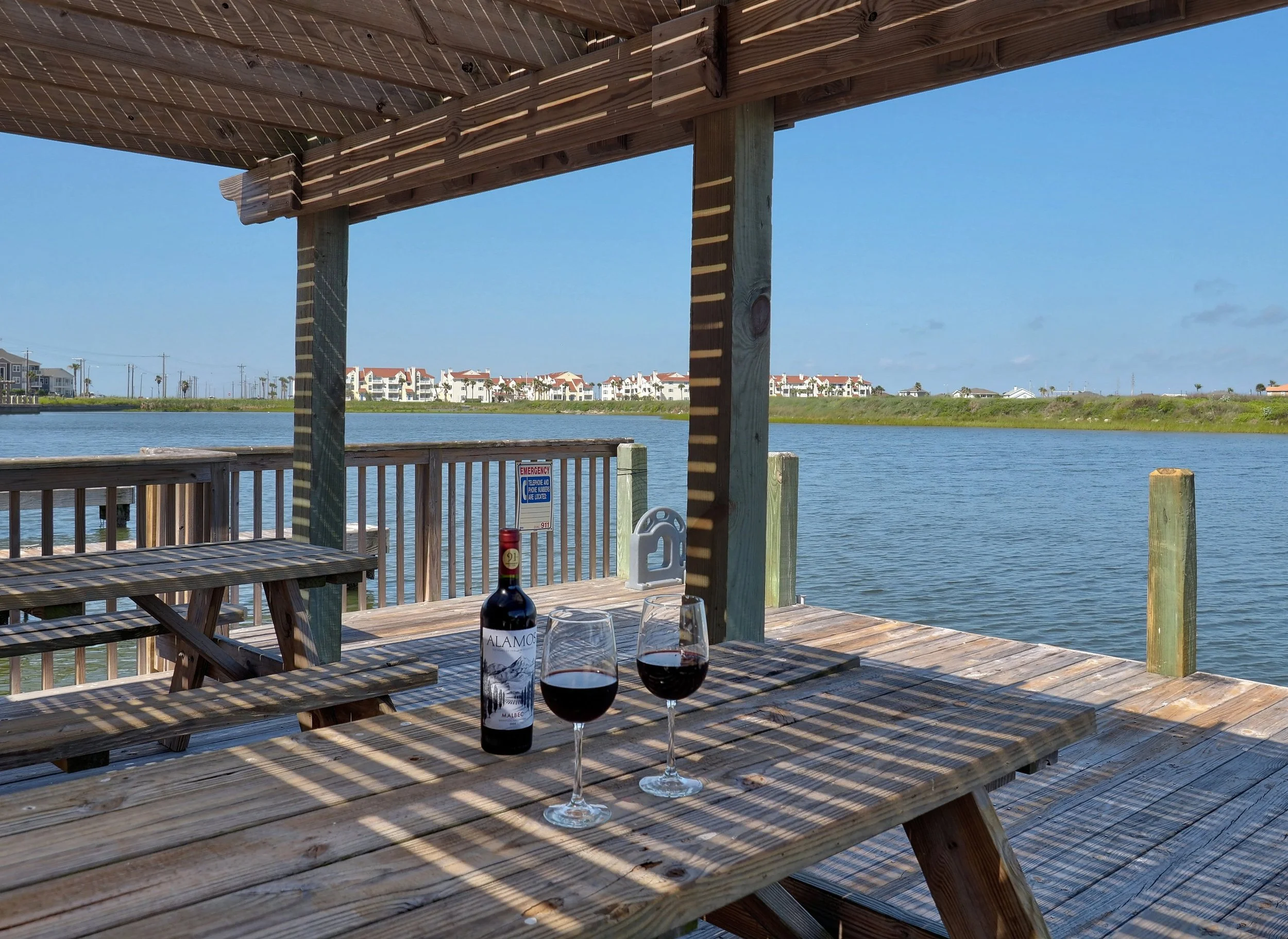 A wooden deck with picnic tables overlooking a body of water. Two glasses of red wine and a bottle of wine are on the table. The scene is sunny with shadows cast by the wooden structure above, and in the background, there are townhouses and a clear b