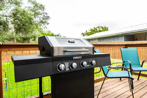 Outdoor patio with a black propane grill and two blue chairs on a wooden deck, green trees in background.