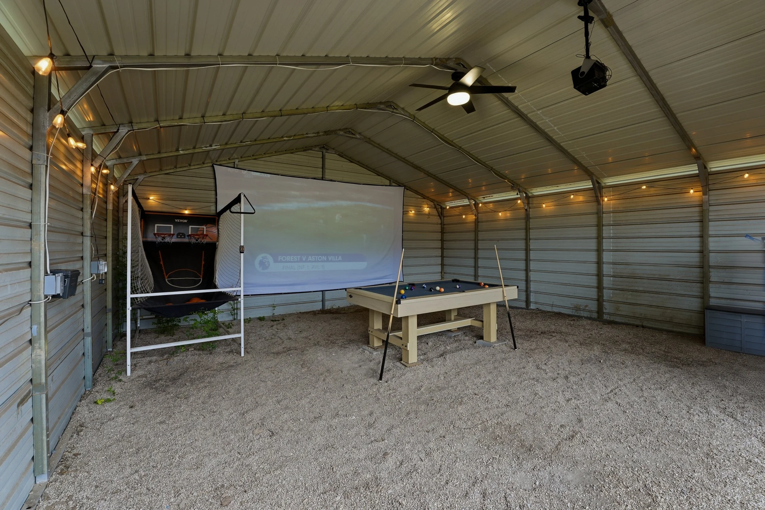 An indoor recreational area with a projector screen displaying a soccer match, a small basketball hoop with ball, a pool table, ceiling fan, string lights, and a speaker system, all within a metal-roofed structure with gravel flooring.