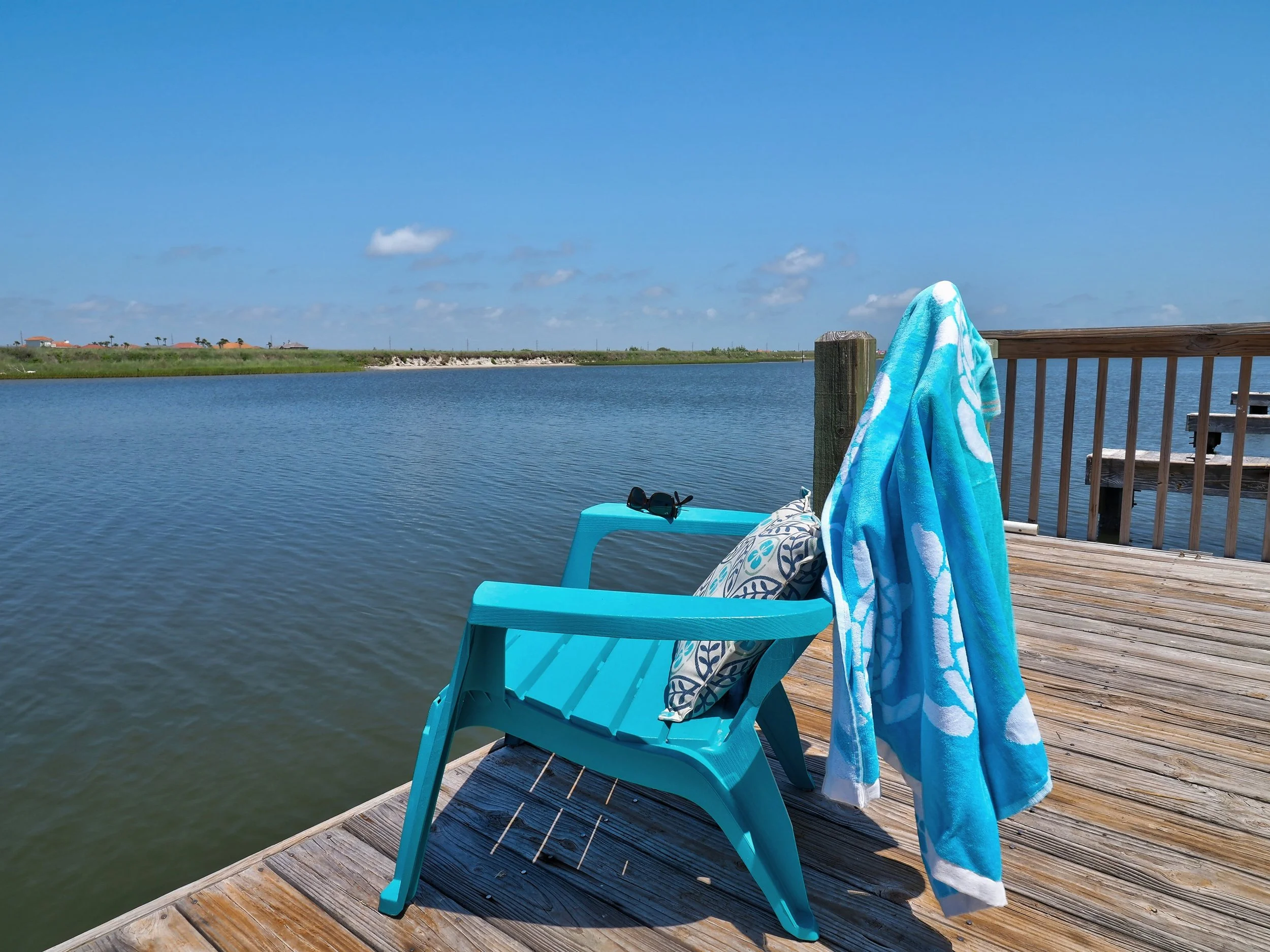 View of a balcony over a body of water with a blue sky and scattered clouds. On the balcony, there is a blue chair with sunglasses on it, a patterned pillow, a blue towel hanging over the railing, and a wooden deck.