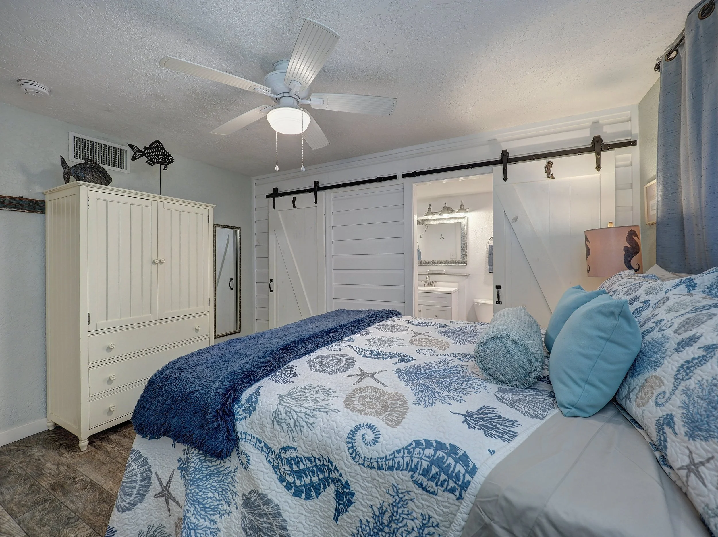 A bedroom with a white bedspread decorated with blue coral patterns, blue pillows, a white cabinet, and nautical decor including a fish sculpture and a metal fish wall art, with a sliding barn door leading to a bathroom.