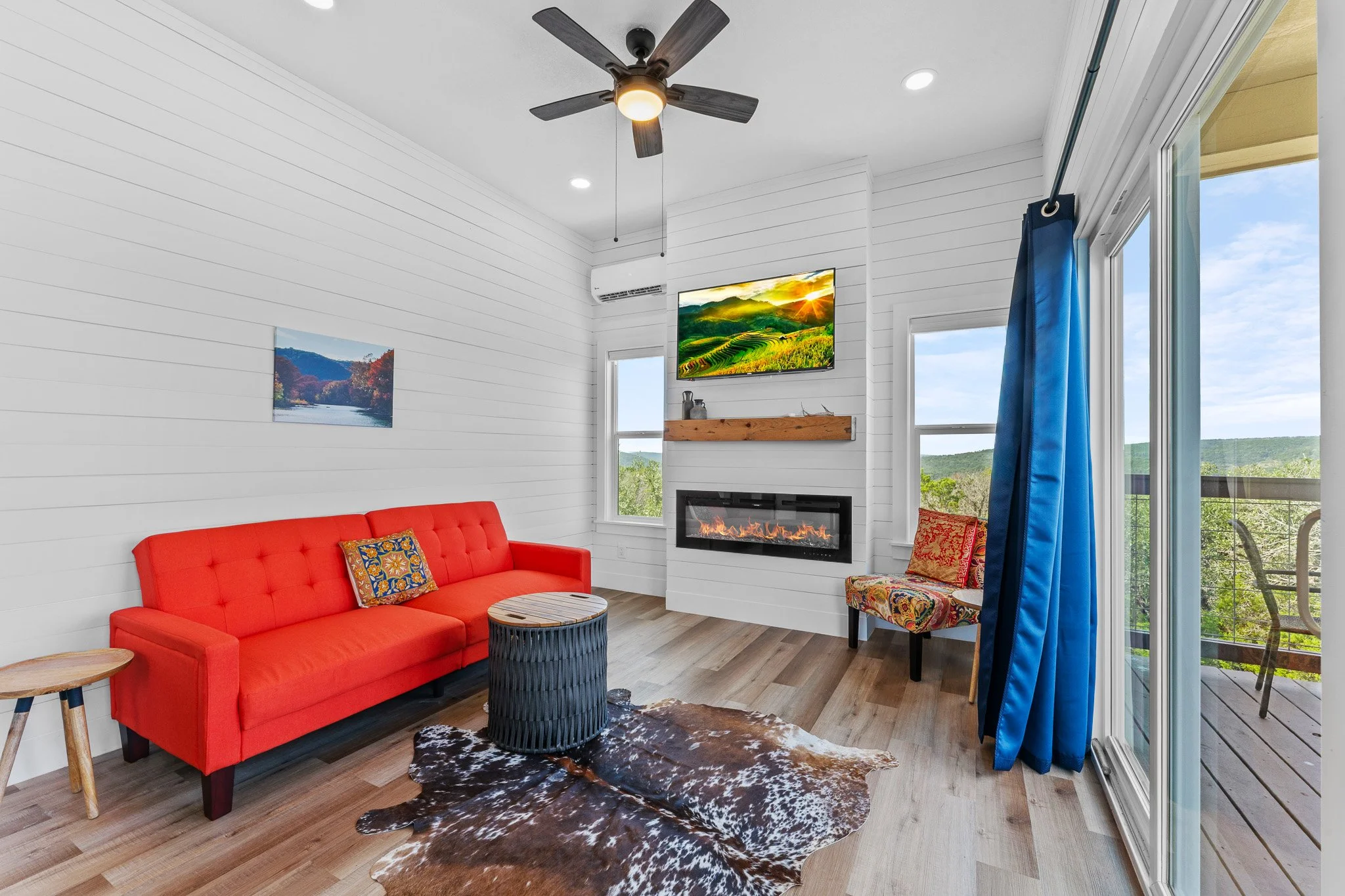 A living room featuring a red sofa with colorful cushions, a cowhide rug, a small round wooden coffee table, a wall-mounted TV displaying a landscape, a fireplace, and large windows with blue curtains showing a scenic view of green hills and a deck o