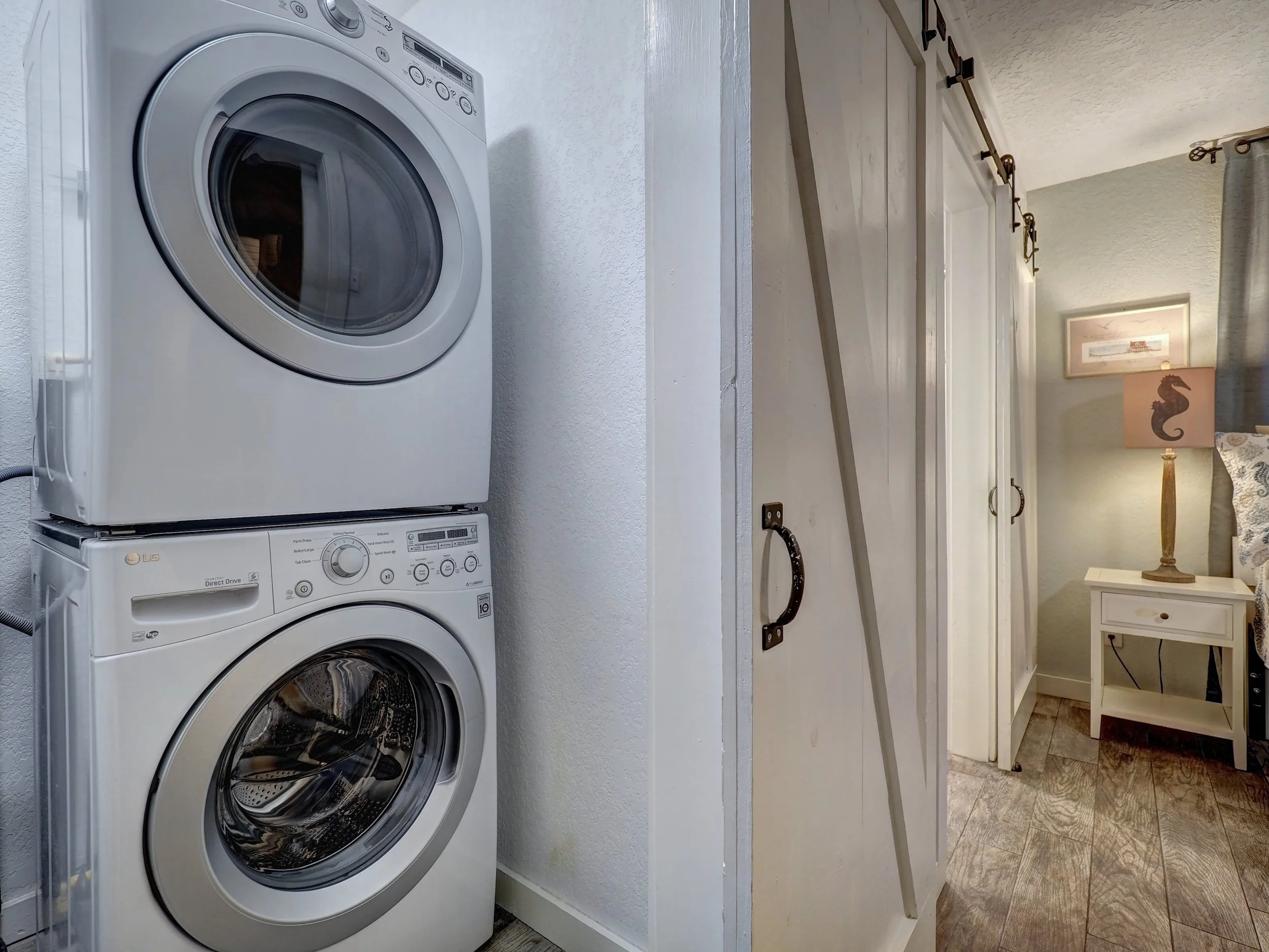 A bathroom with a stacked washer and dryer, a white countertop, a wooden cabinet with black handles, and a bedside table with a lamp featuring a pink shade with a seahorse design.