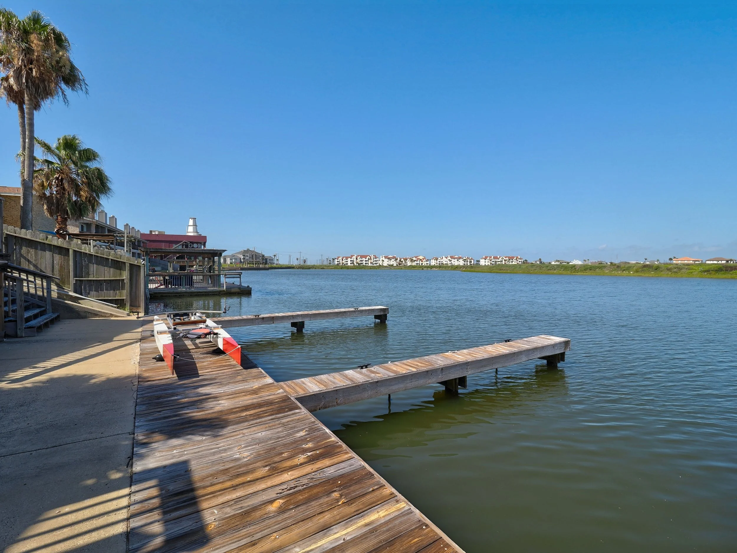 A waterfront scene with a dock extending into a calm body of water, shaded by palm trees on the left, with residential buildings in the background under a clear blue sky.