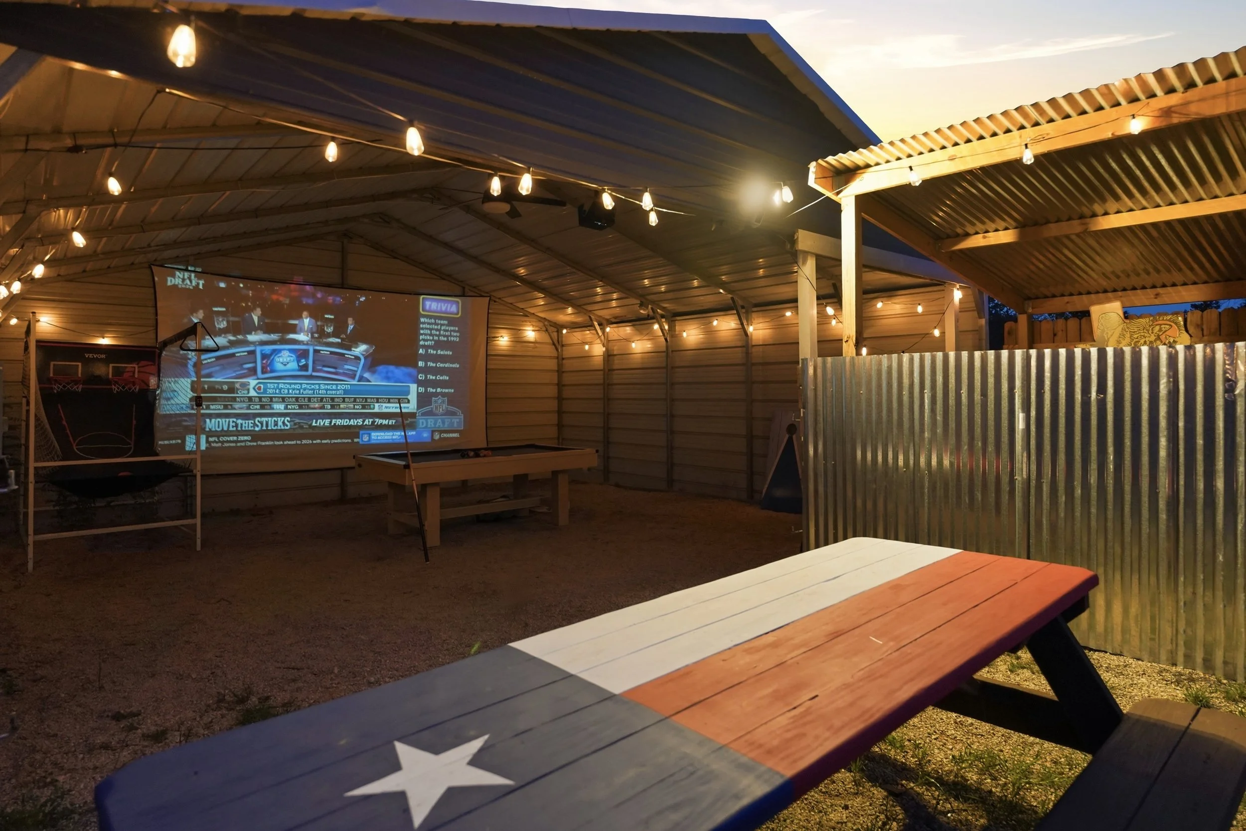 Outdoor covered patio with string lights hanging from the ceiling. A large projection screen displays a sports trivia game. There are wooden picnic tables, one painted with the American flag design, and a corrugated metal divider partially visible on