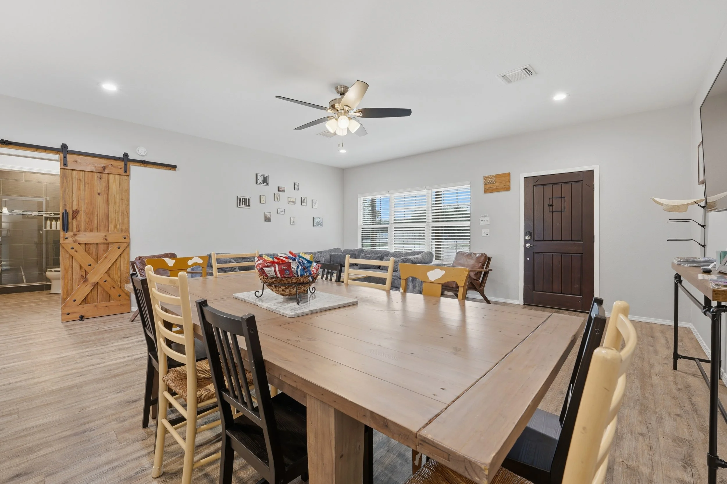 A dining area with a wooden dining table, surrounded by black and cream chairs, in front of a distressed gray wall with photo frames. There is a window with white blinds, a ceiling fan, and a sliding barn door leading to a bathroom.