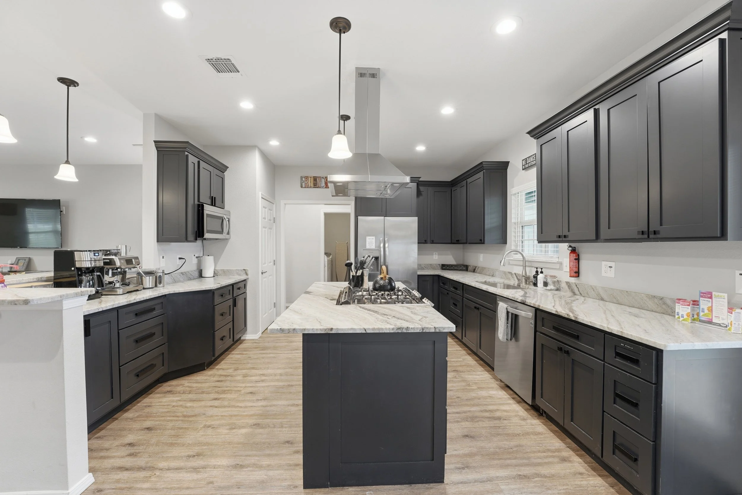 Modern kitchen with dark gray cabinets, granite countertops, wooden flooring, stainless steel appliances, island in the center, and bright ceiling lights.