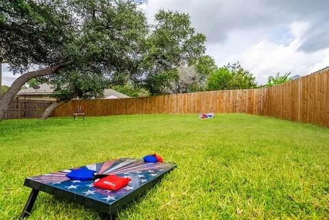 Backyard with green grass, a wooden fence, and outdoor game pieces including a cornhole board with red and blue bags, some toys in the distance, and a tree under a cloudy sky.