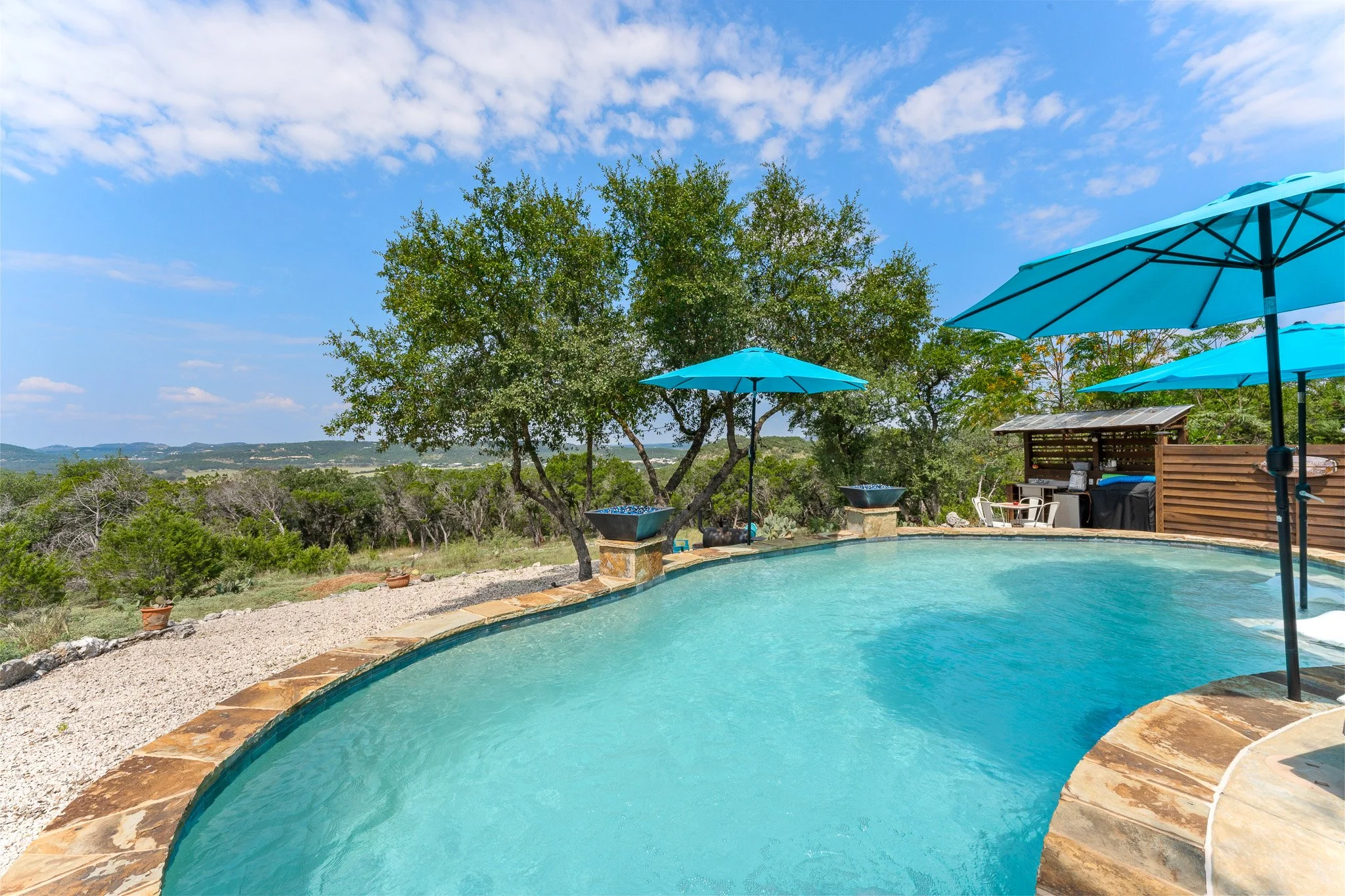 An outdoor swimming pool with a stone border, blue umbrellas, and a shaded sitting area, set against a backdrop of green trees and a distant view of rolling hills under a partly cloudy sky.