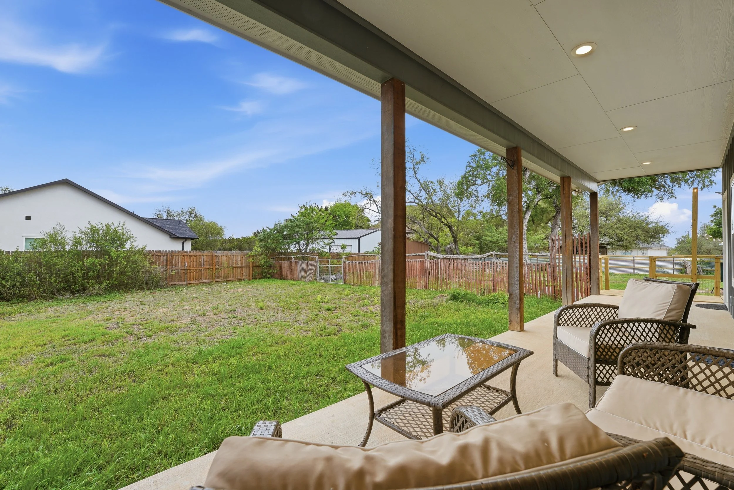 A backyard patio with outdoor furniture, including cushioned chairs and a glass-top table, overlooking a green yard enclosed by a wooden fence with neighboring houses and trees in the background under a blue sky.