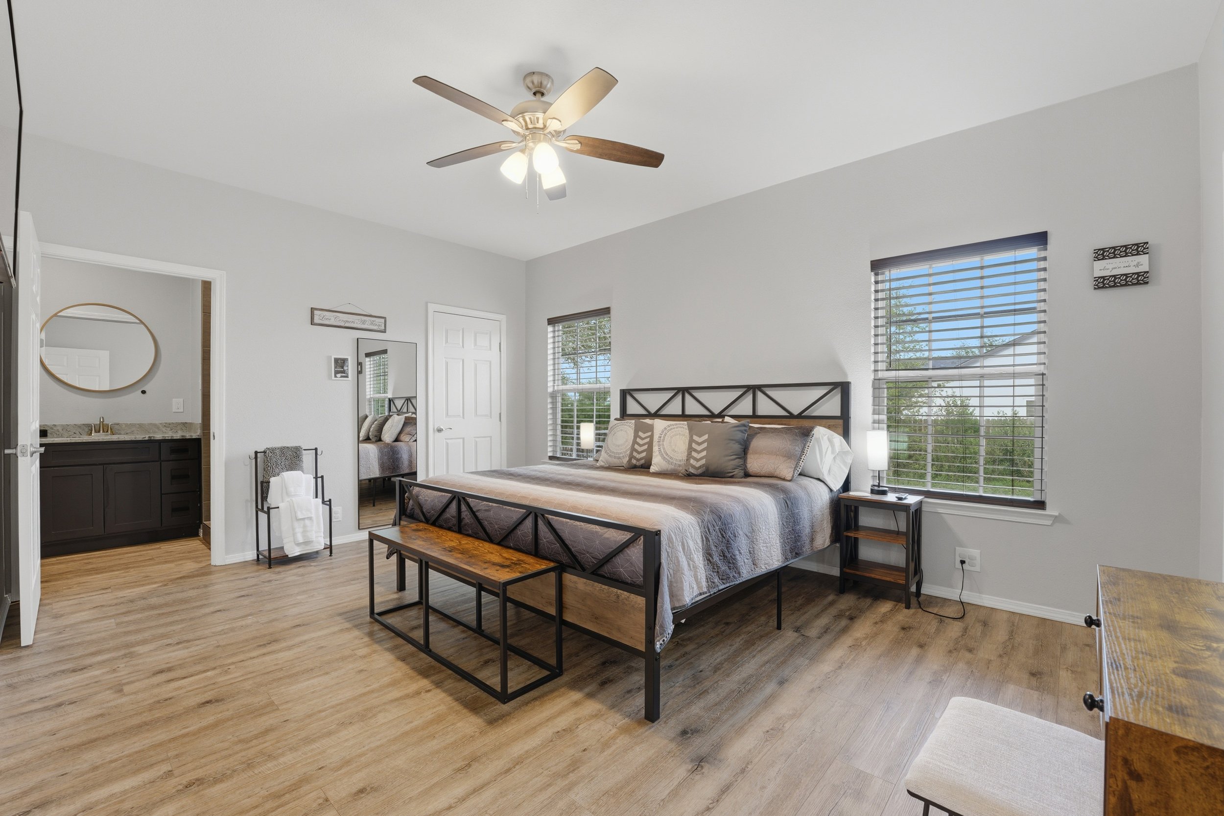 Bright bedroom with large windows, a double bed with patterned pillows, a ceiling fan, and a wooden bench at the foot of the bed. There is a nightstand with a lamp and a small mirror, and an open door leading to a bathroom.