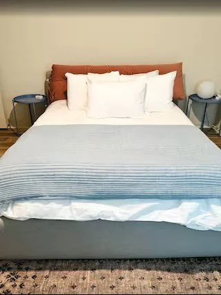 A neatly made bed with a brown headboard, white pillows, and a gray and white striped blanket in a bedroom with two dark blue nightstands and white lamps.