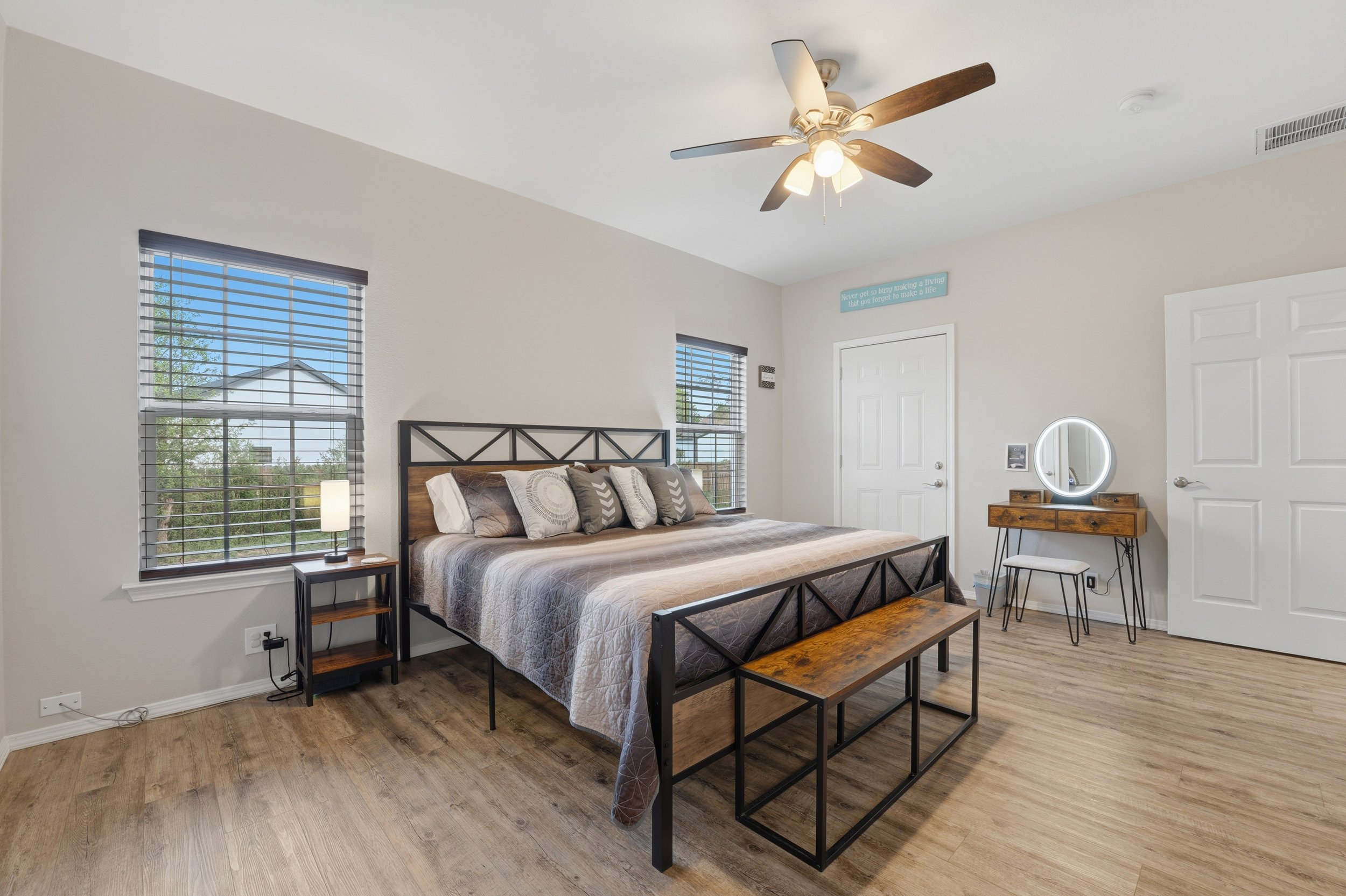 A bedroom with a bed, two nightstands, a vanity with a mirror, and two large windows with blinds.