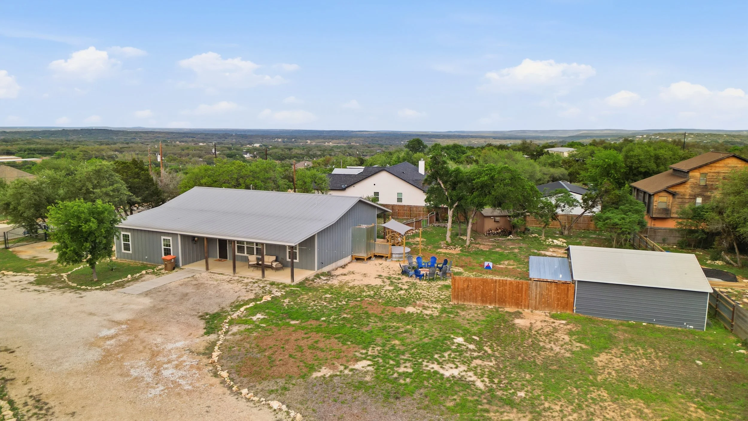 A backyard with a gray house, outdoor seating, a wooden deck, trees, a fenced yard, and neighboring houses, with a view of distant rolling hills under a partly cloudy sky.