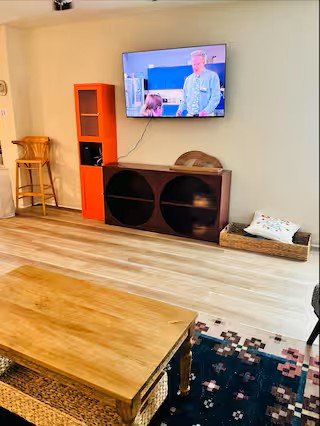Living room with a wall-mounted TV showing a man speaking, orange bookshelf, wooden sideboard, yellow chair, woven basket with a pillow, wooden coffee table, and a patterned rug on light laminate flooring.