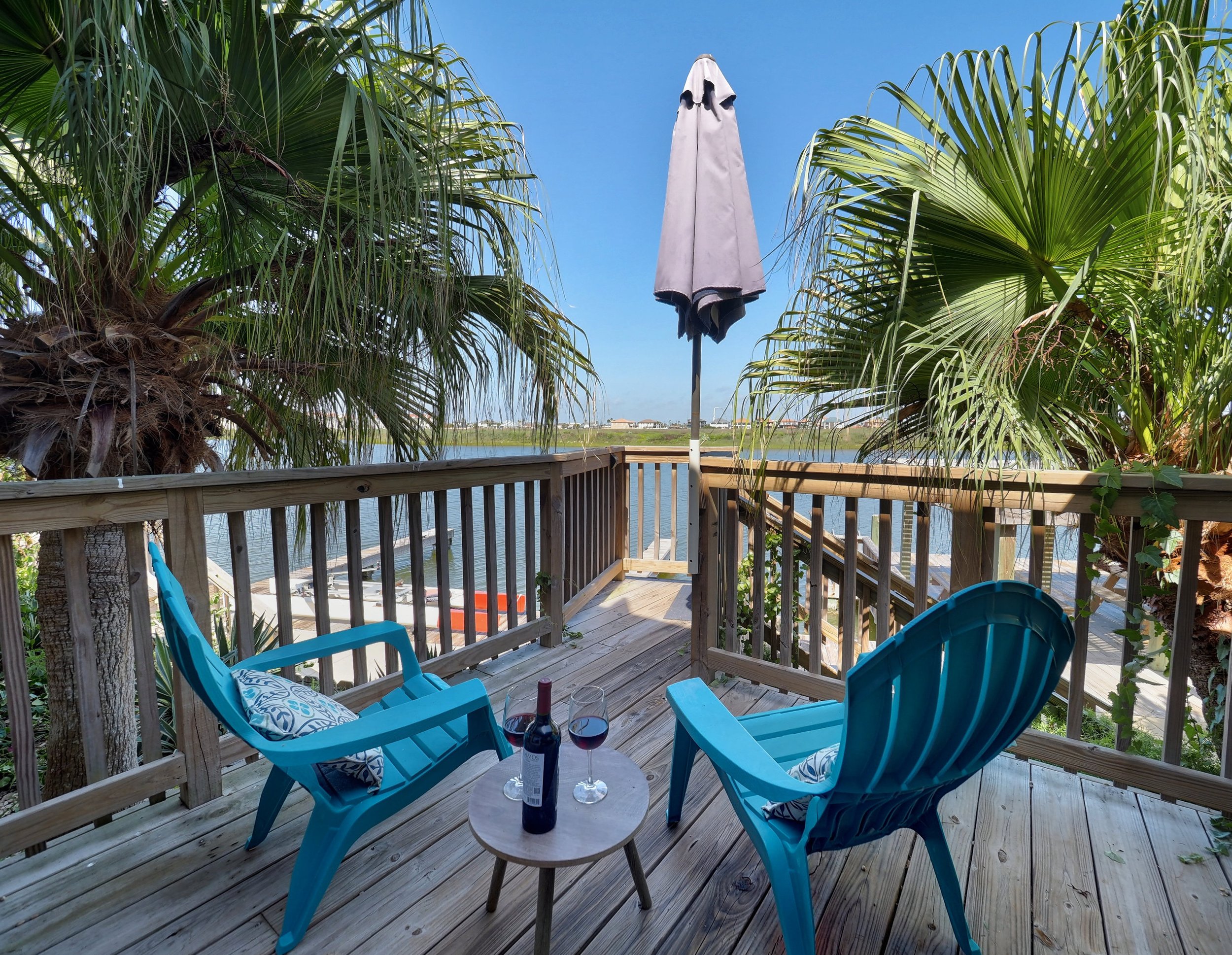 View from a wooden deck with two blue chairs and a small round table with two glasses of red wine and a bottle, overlooking a body of water with houses on the opposite shore. A closed patio umbrella stands between two lush green palm trees, with a cl