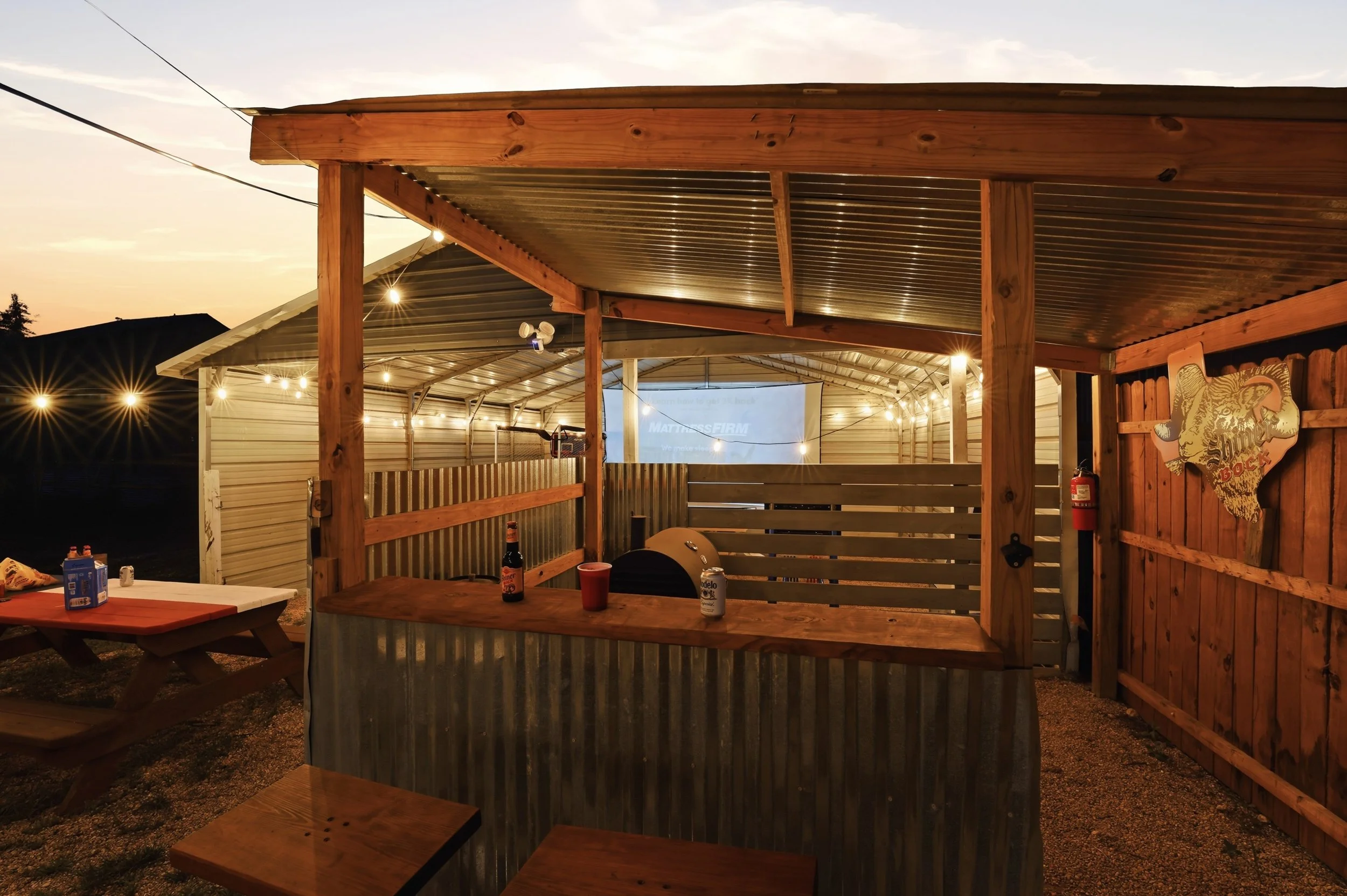 Outdoor patio area during sunset with string lights, a wooden picnic table with drink bottles, a corrugated metal and wood structure, a fire extinguisher, and a rustic sign with a tiger on the wooden fence.