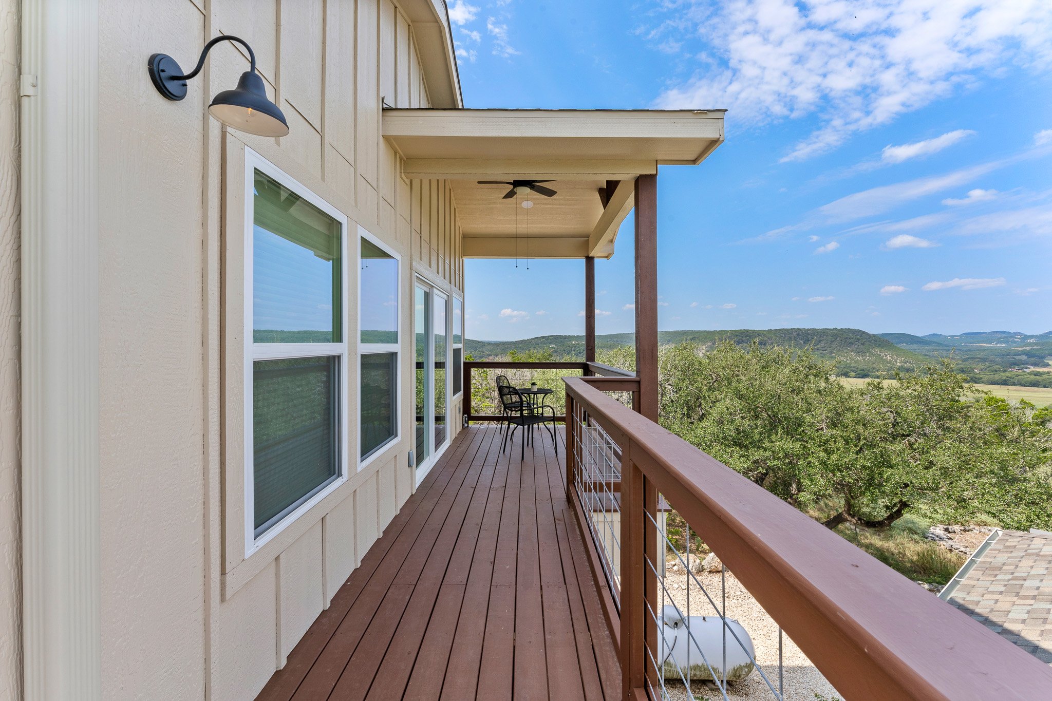 A balcony with wooden flooring, railing, and a ceiling fan, overlooking a green landscape with trees and mountains in the distance under a partly cloudy blue sky.