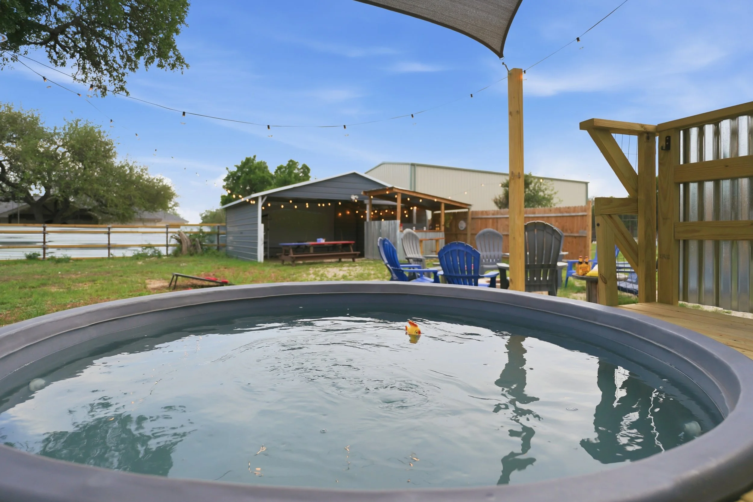 Backyard with a small hot tub in the foreground, a wooden deck, plastic chairs, a shed with string lights, and a fenced yard with trees and a blue sky.