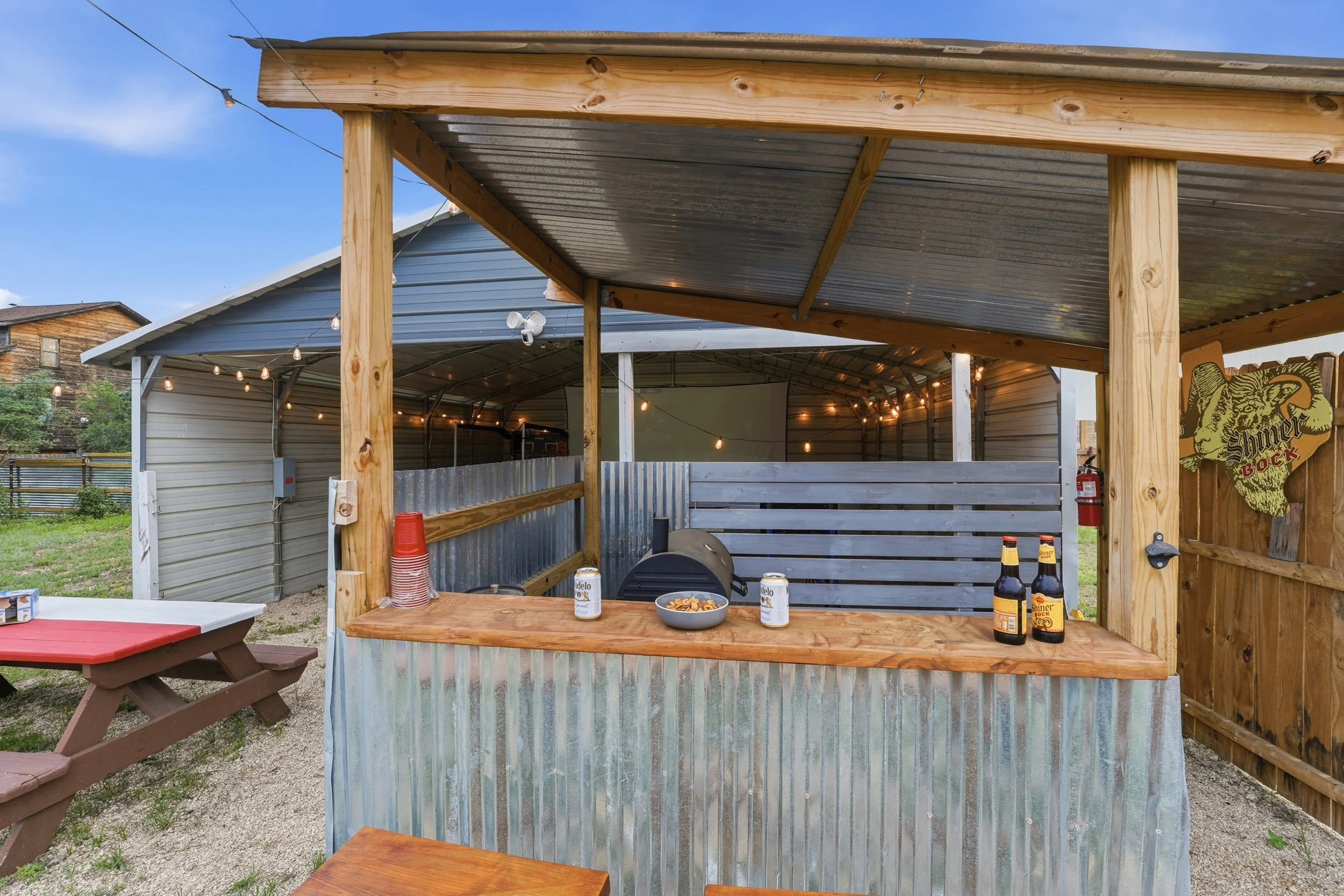 A covered outdoor bar area with a wooden countertop, bottles of Shiner Bock beer, a bowl of snacks, and cans of Modelo beer. There are string lights hanging overhead and part of a picnic table with a red and brown surface in the foreground. The backg