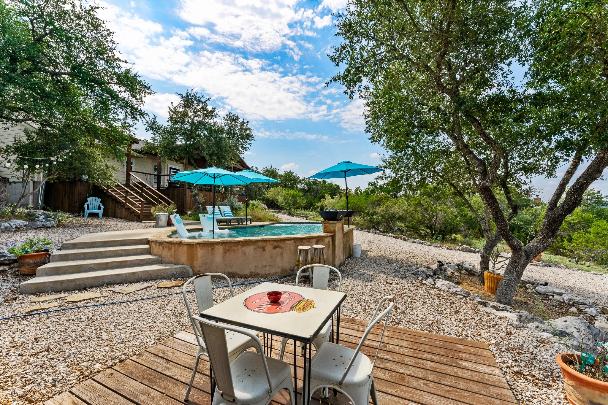 A backyard scene with a swimming pool, outdoor seating including white chairs and a small table, large blue umbrellas, a house with stairs, trees, and a partly cloudy sky.