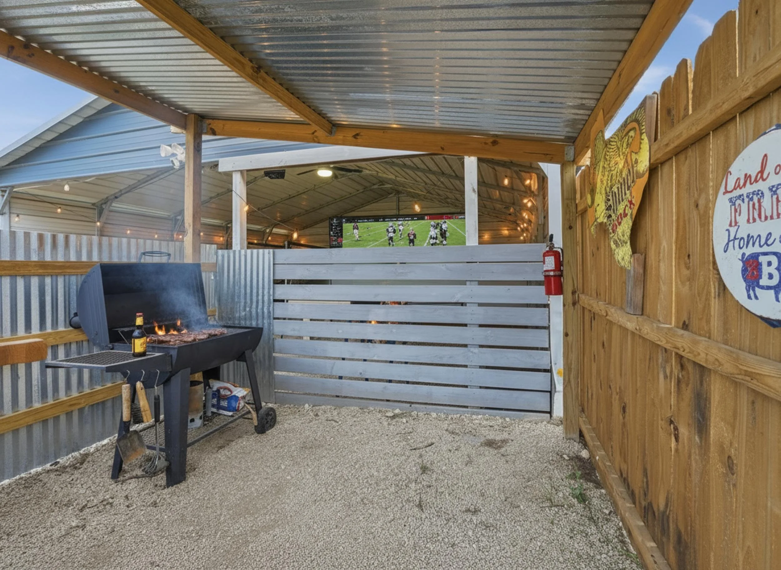 A covered outdoor area with a grill, a wooden fence, rustic signs, and string lights, overlooking a yard with a large screen displaying a football game.