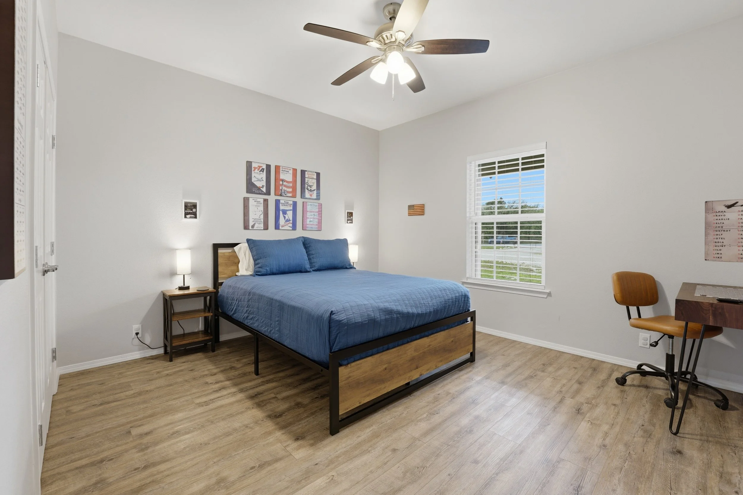 A bedroom with a bed covered in a blue quilt and pillows, a small wooden nightstand with a lamp on each side, wall art above the bed, a window with white blinds letting in natural light, and a wooden chair near a small wooden desk.