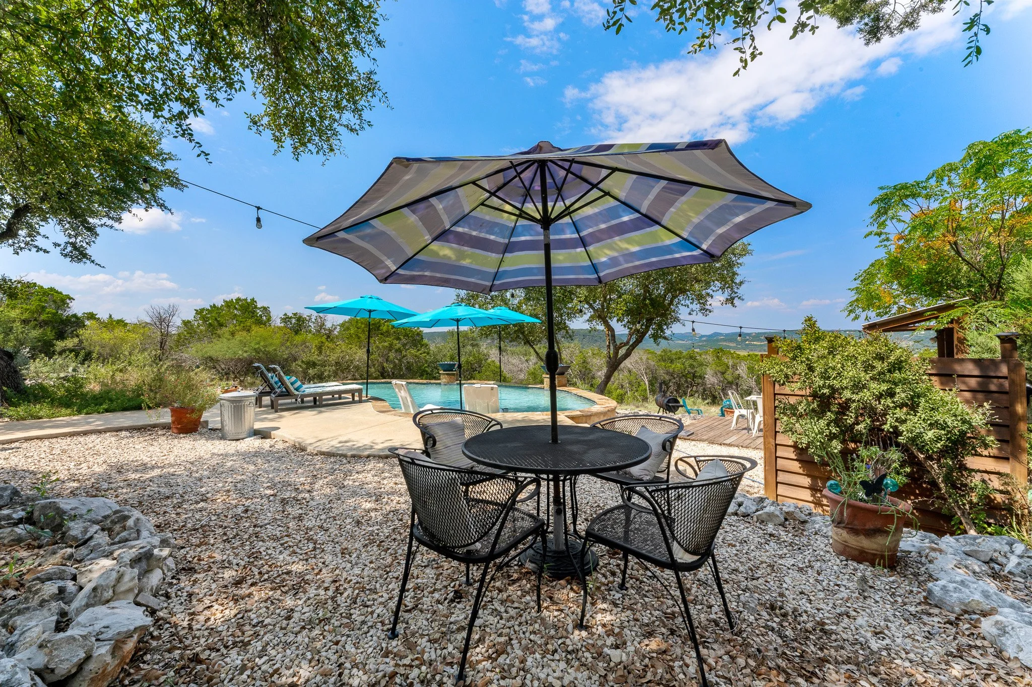 Outdoor pool area with patio furniture and large umbrellas, surrounded by trees and greenery, under a partly cloudy sky.
