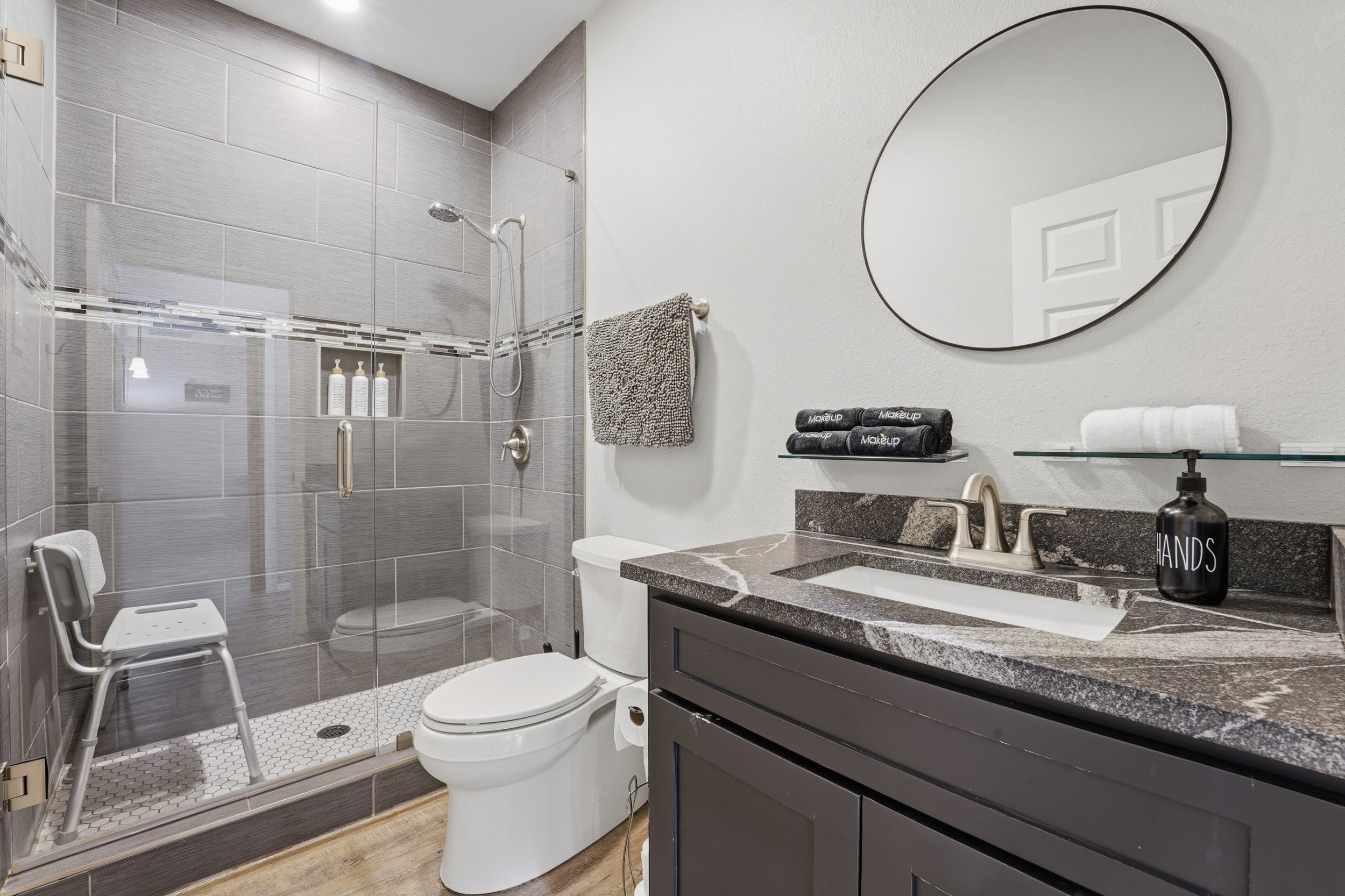 Bathroom with a glass shower enclosure, a toilet, a black vanity with a marble countertop, a round mirror, and various toiletries and towels.